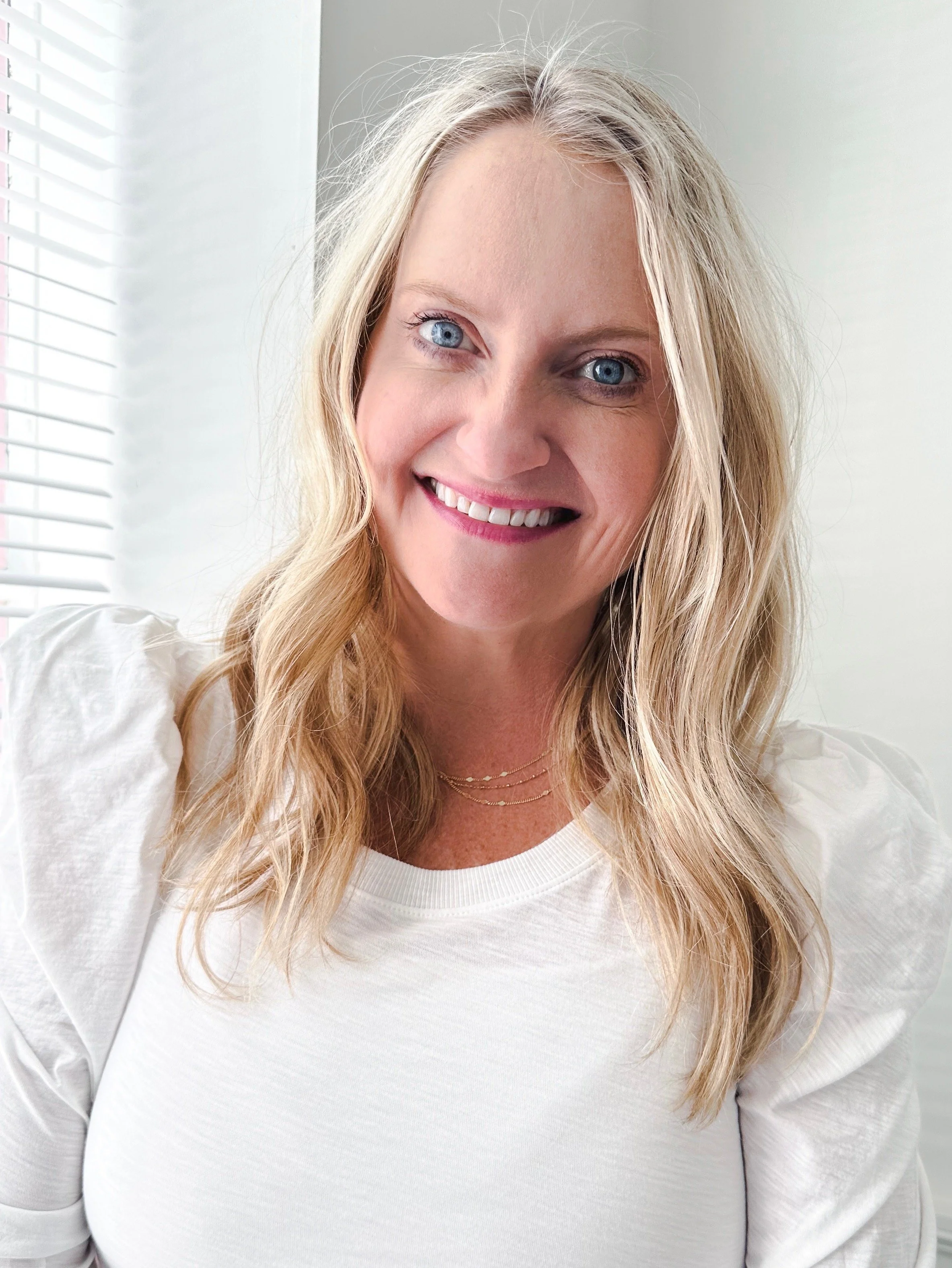 Close-up of a female therapist with long blonde hair, smiling, wearing a white top, indoors with a window in the background.