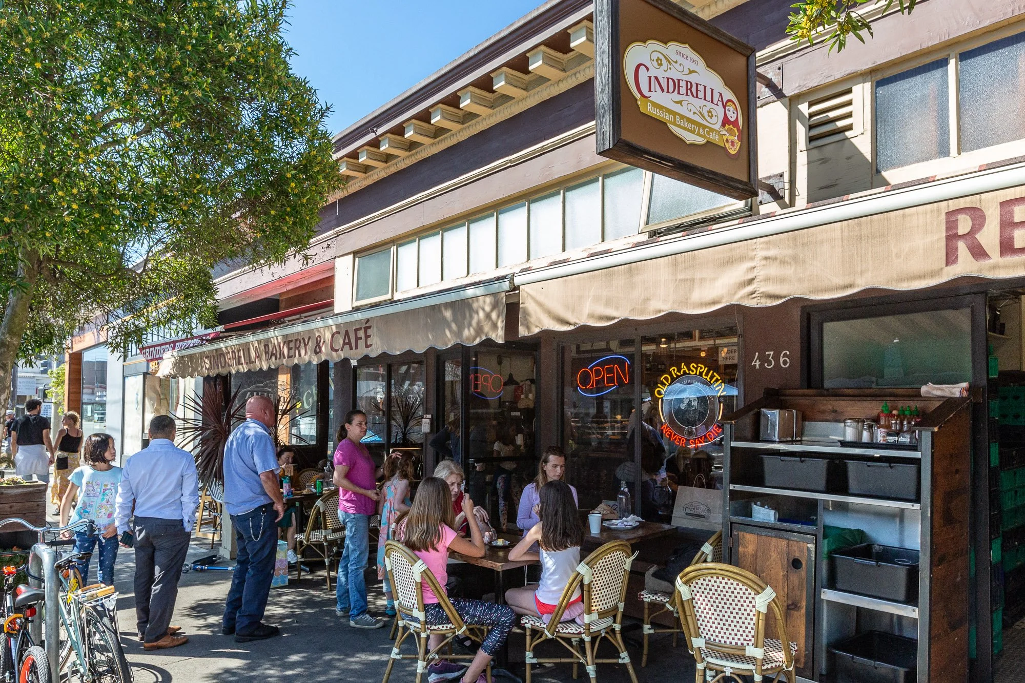 People sitting outside and standing in front of Cinderella Bakery & Cafe, a Russian bakery and café, with an outdoor seating area, neon open sign, and signage indicating it is open.