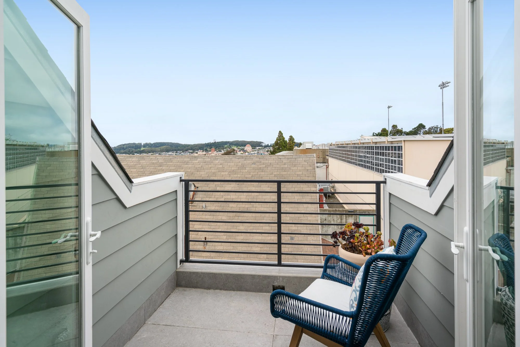 Small balcony with a blue wicker chair and a potted plant, overlooking neighboring rooftops and distant hills under a clear blue sky.