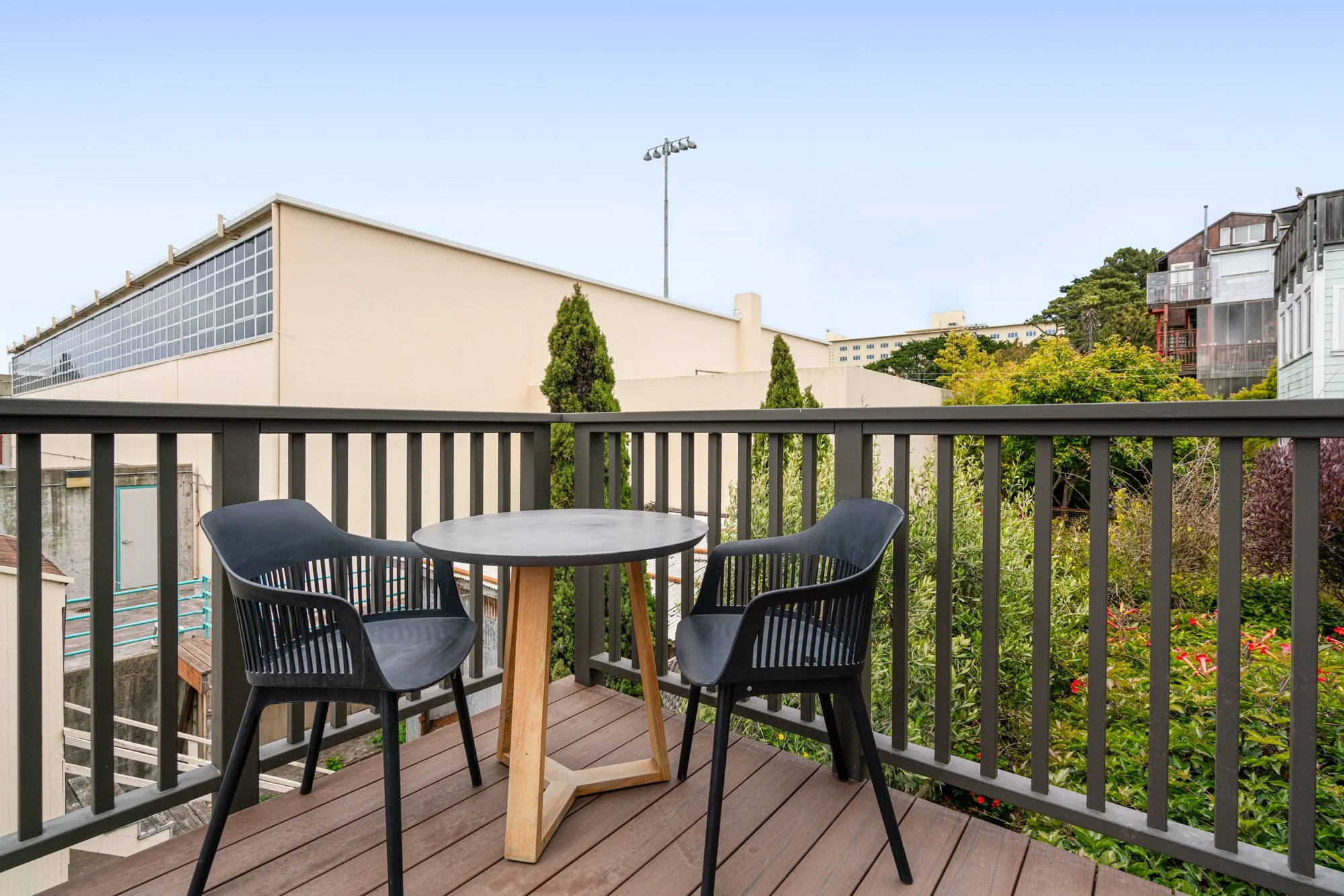 Balcony with a round black table and two black chairs, overlooking a cityscape with buildings and greenery.