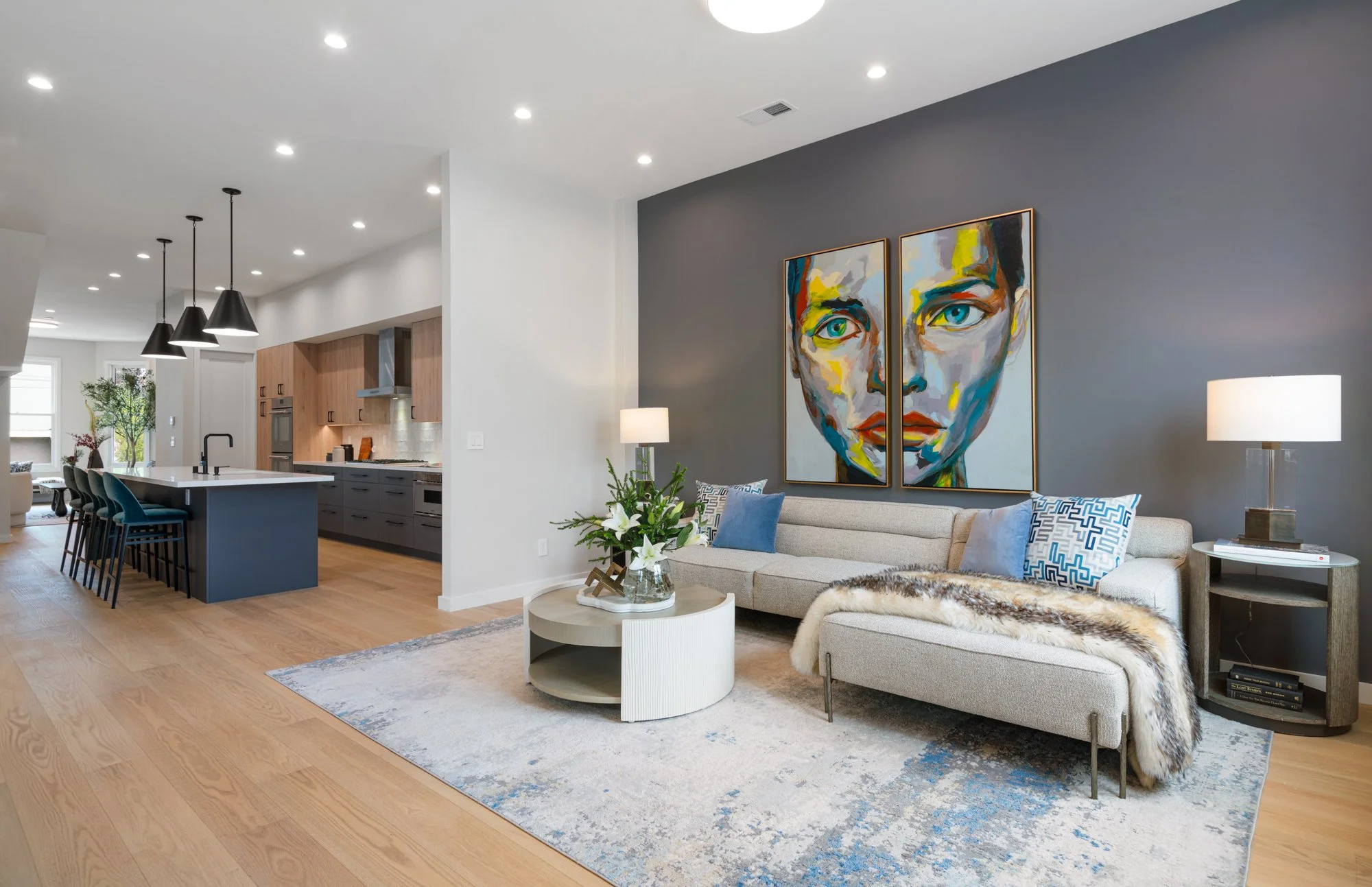 Modern living room with beige sofa, colorful abstract portrait art on dark gray wall, white coffee table, table lamps, and a blue and white rug, open to a kitchen with wooden cabinets, black accents, and black pendant lights.