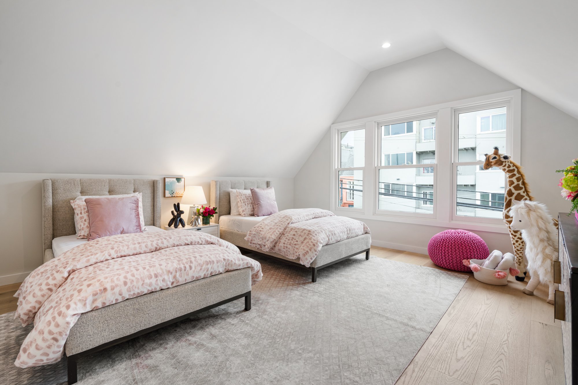 A bright, modern bedroom with two twin beds featuring pink leopard print bedding, a large window with a city view, and stuffed animal giraffe and horse near a pink pouf and a white basket with plush slippers.