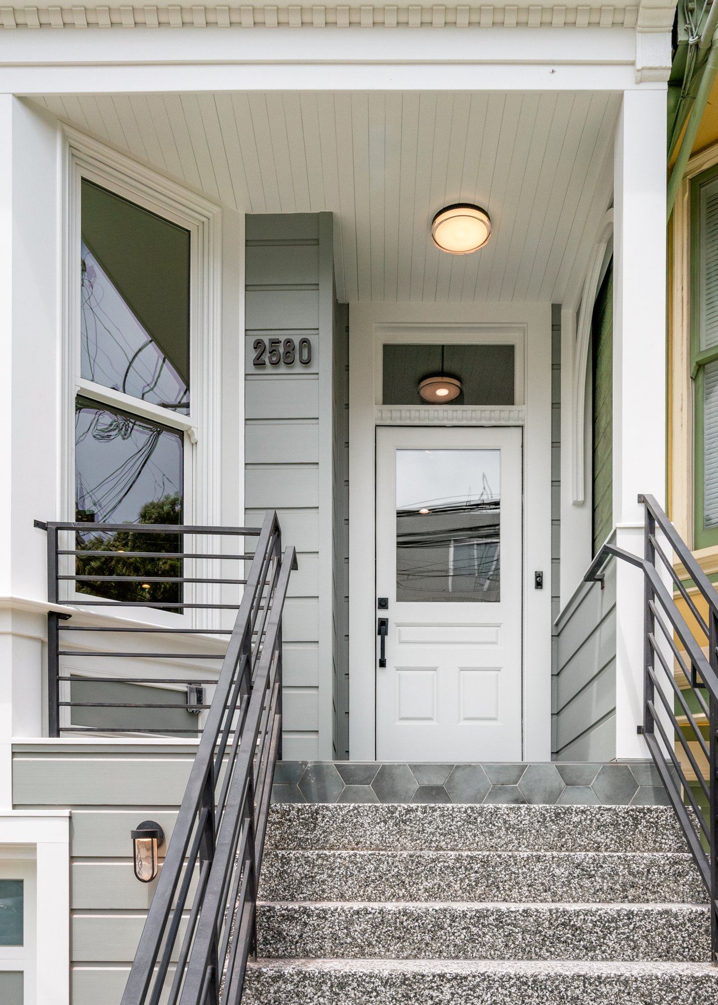 Front porch of a house with gray and white siding, black metal railings, a concrete stoop, and a white door with a glass window, displaying the house number 2580.