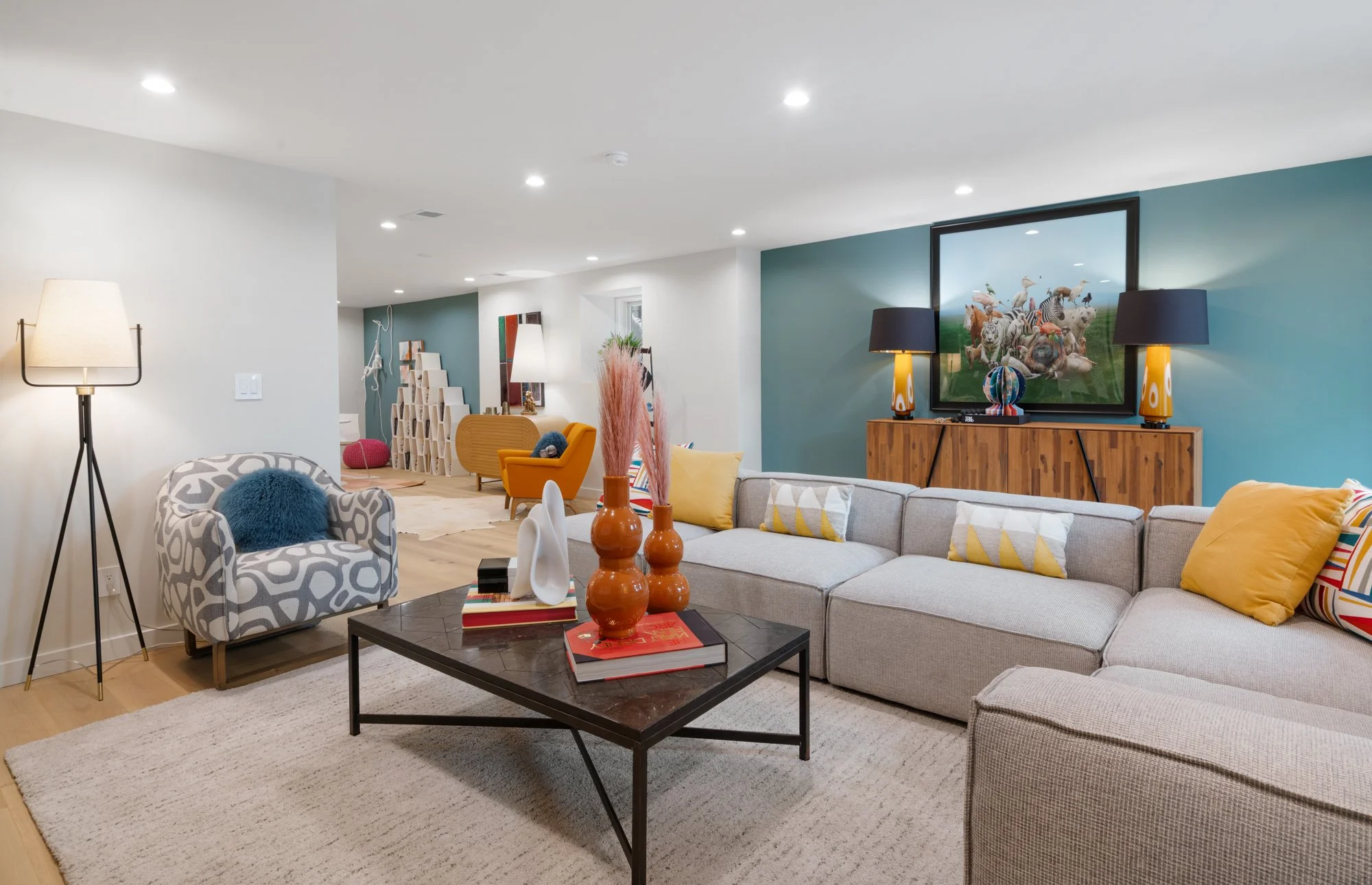 Living room with beige sectional sofa, colorful pillows, a black coffee table with vases and books, a patterned armchair with a blue pillow, a wooden sideboard with a TV and lamps, hardwood floors, white walls with accent walls, and decorative items 