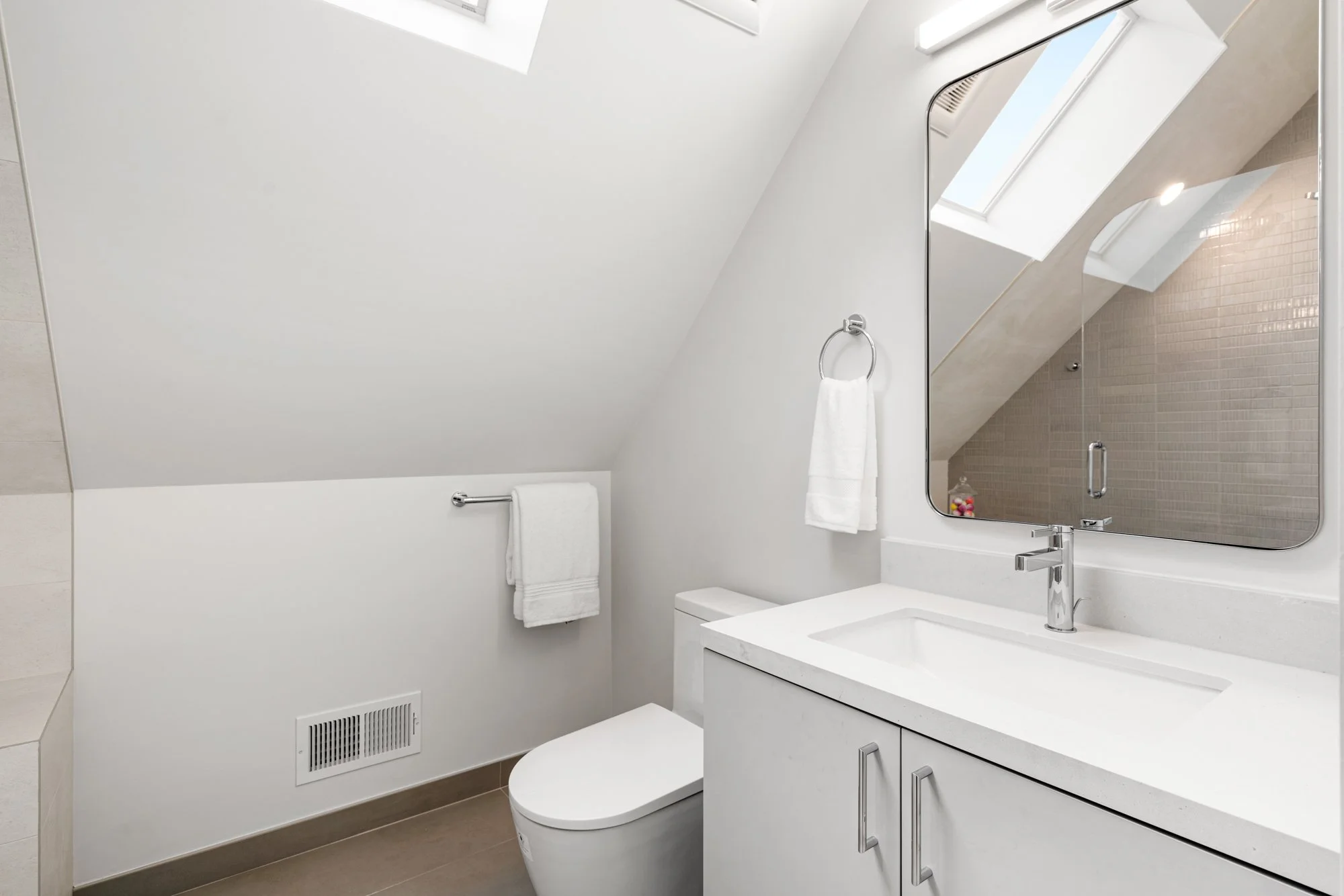 A modern, minimalist bathroom with white walls and a sloped ceiling, featuring a white toilet, a white vanity with a built-in sink, a large mirror, and a chrome towel ring and towel, illuminated by natural light from skylights.