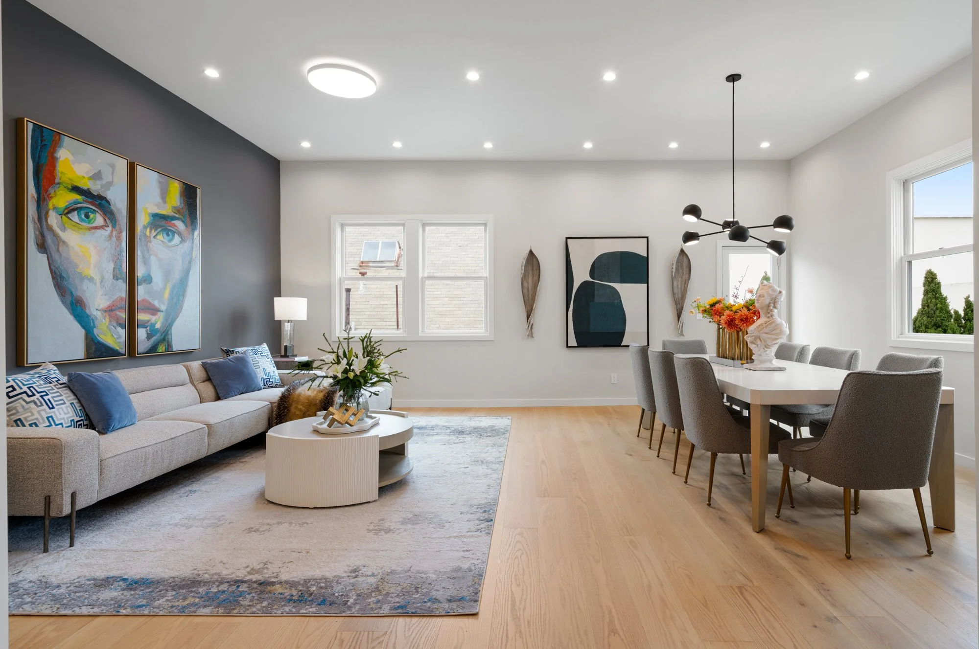 Living room with gray sofa, coffee table, and artwork on the wall, adjacent to a dining area with a table, cushioned chairs, floral centerpiece, and modern chandelier, with hardwood floors and large windows.
