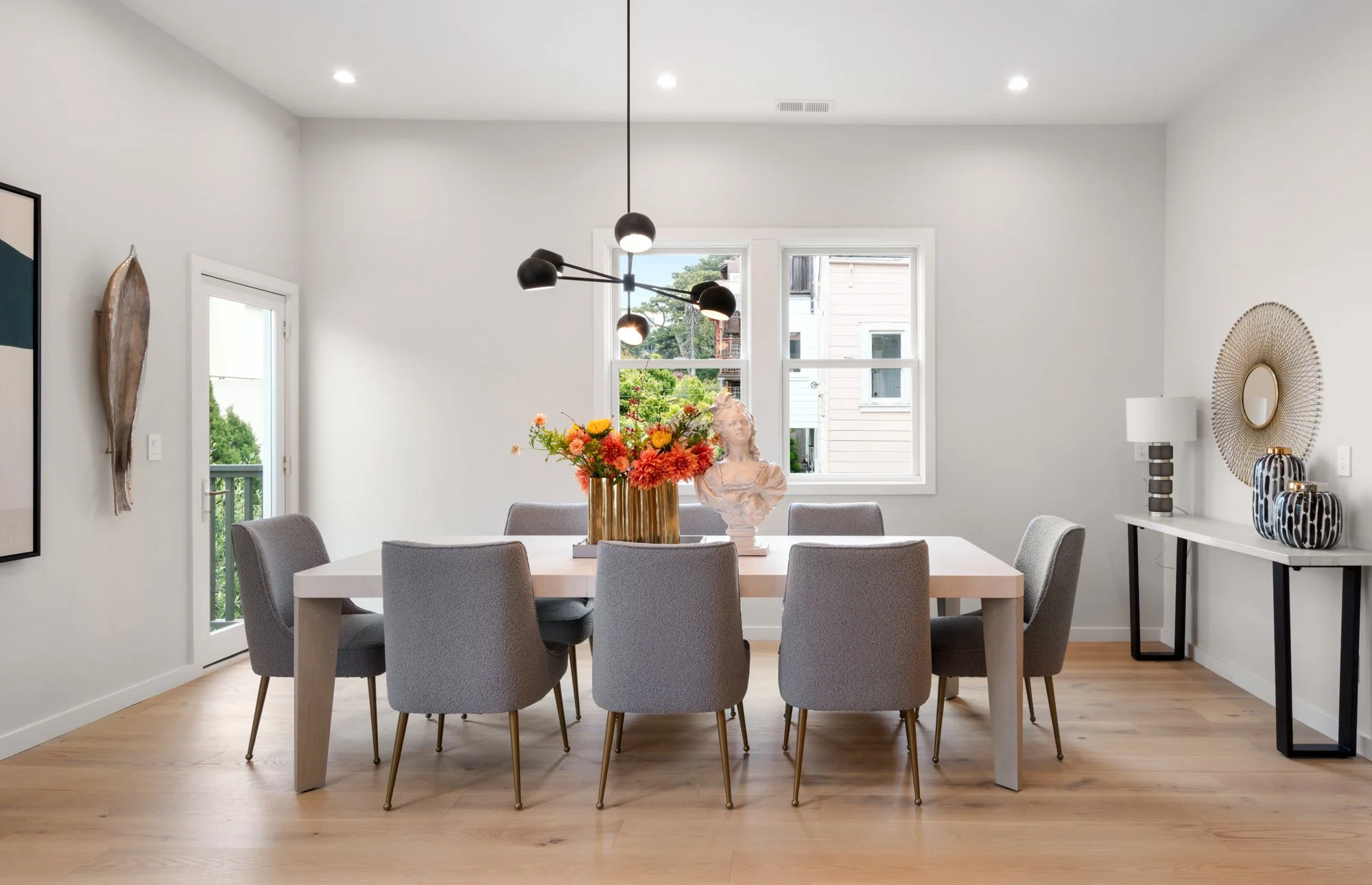 Modern dining room with light wood flooring, gray upholstered chairs, a white dining table, a black multi-bulb pendant light, a large floral centerpiece, a white sculpture, and decorative vases, with a window and wall art.