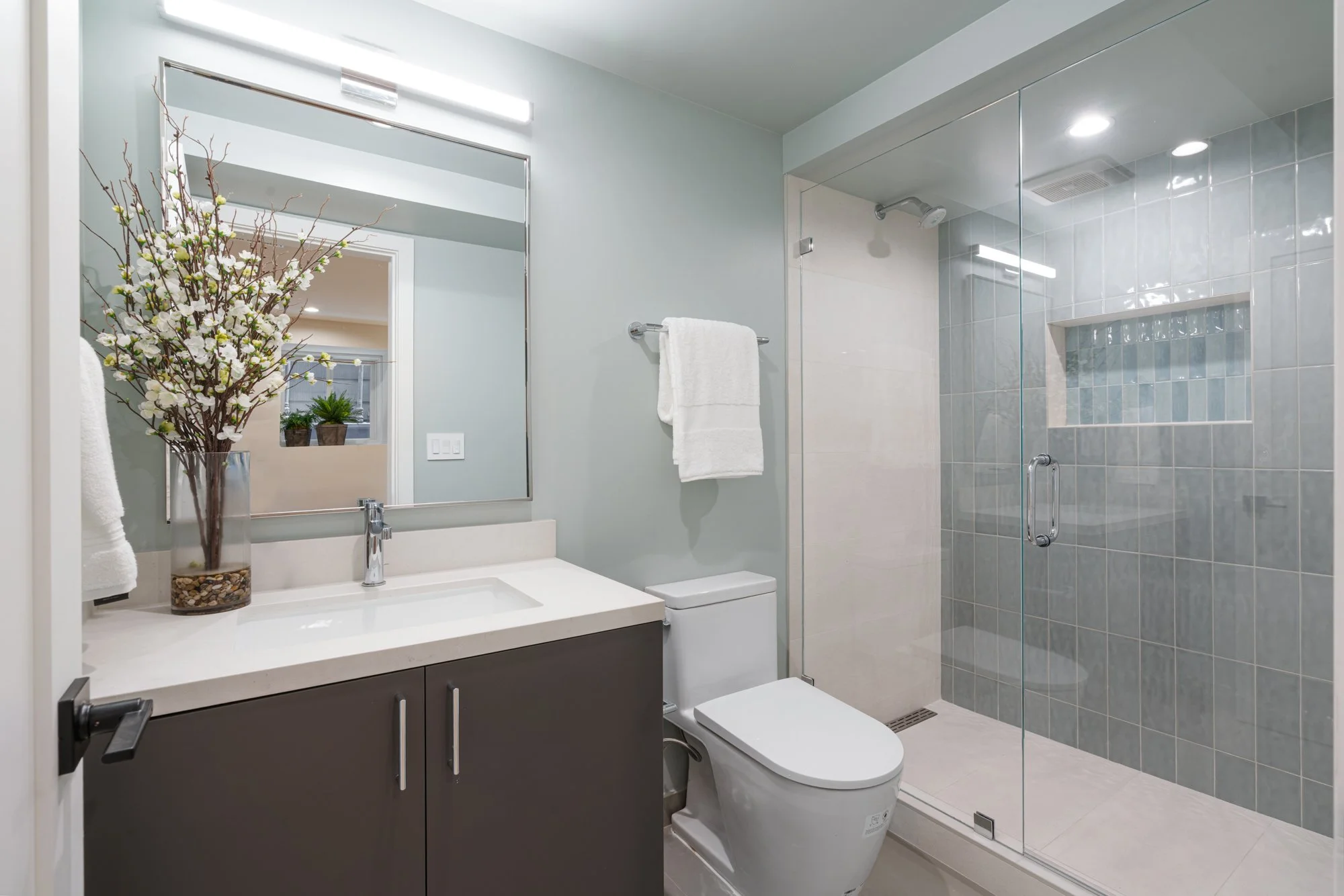 Modern bathroom with glass-enclosed shower, white toilet, vanity with dark cabinets, large mirror, and decorative flower arrangement.