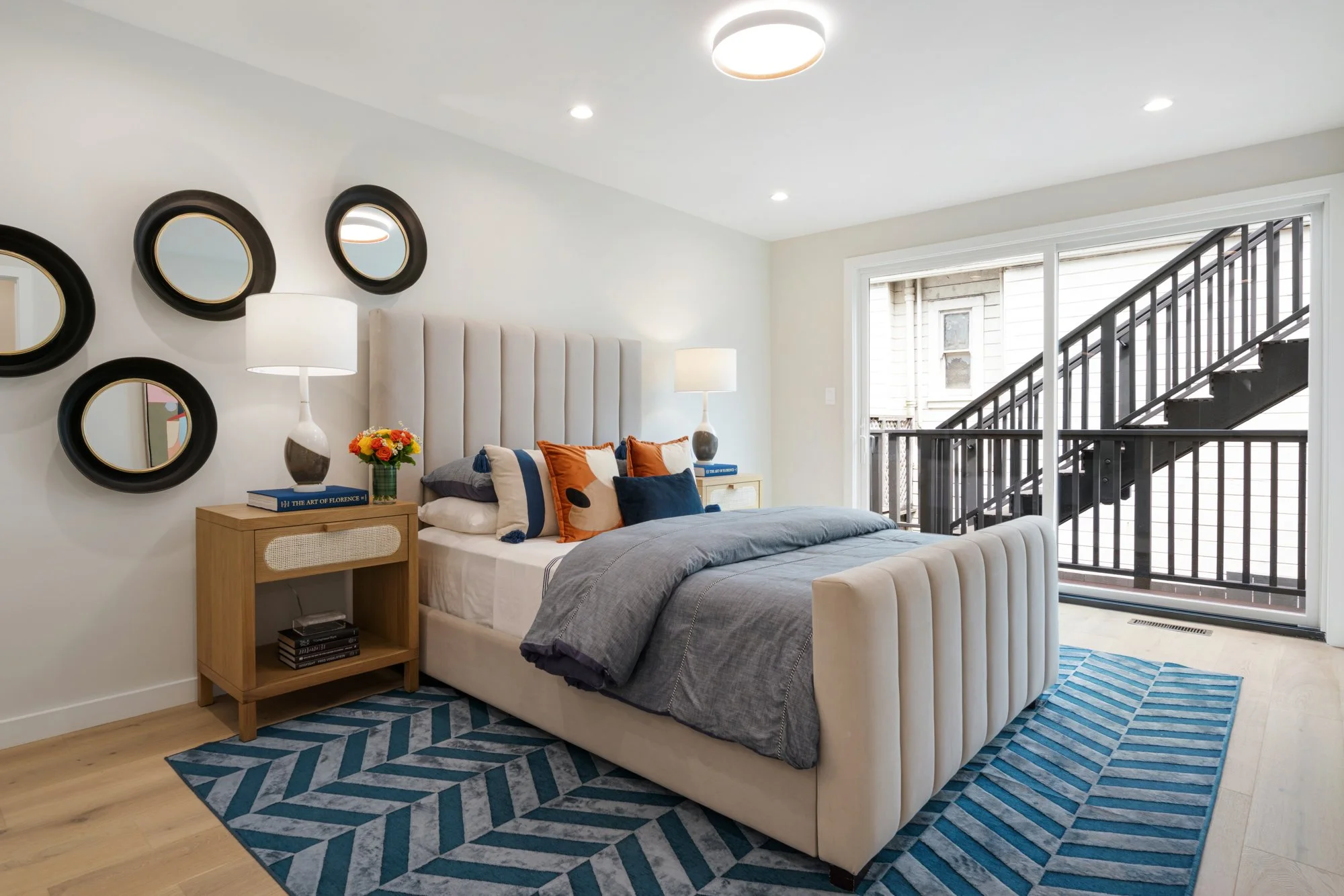 Bedroom with beige upholstered bed, wooden nightstands with lamps, blue and orange accent pillows, striped blue rug, sliding glass door leading to balcony, and wall mirrors.