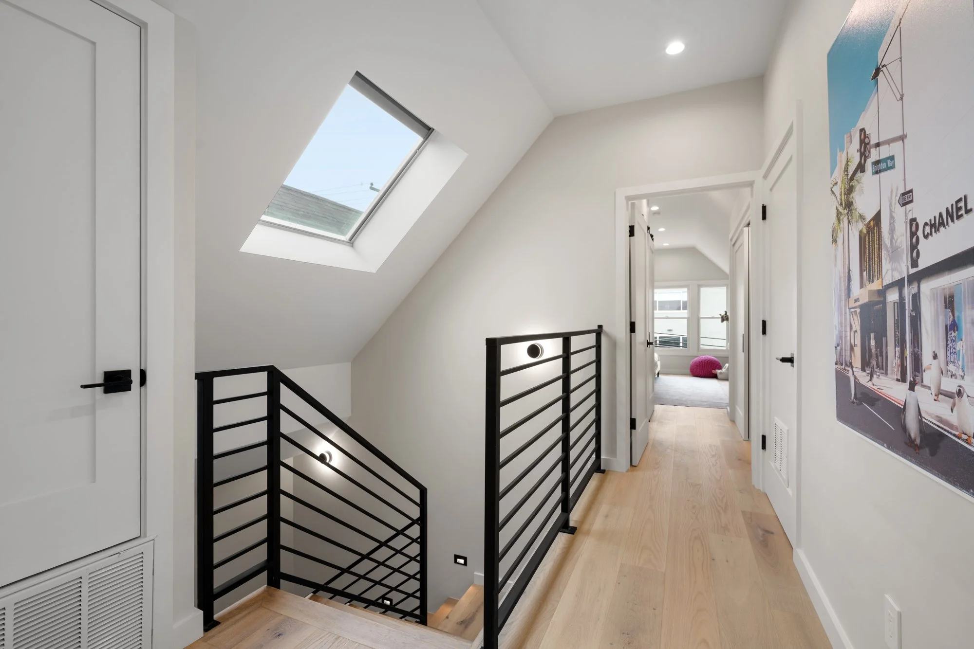 Interior view of a modern house hallway with white walls, a skylight, and a black metal staircase railing leading down. There are four closed white doors on the right side and a large wall poster on the right wall. At the end of the hallway, a bright