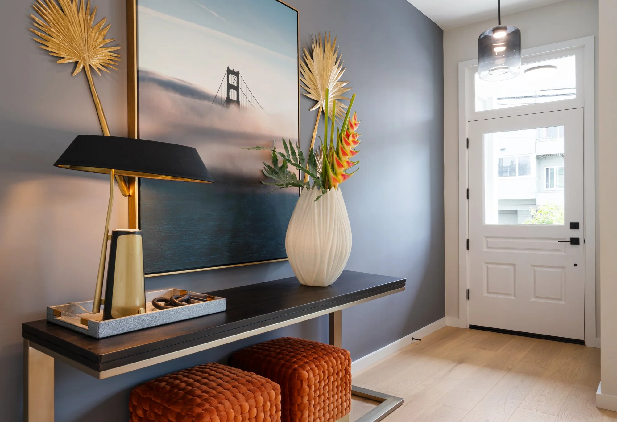 Modern entryway with a black console table, large wall art of a bridge and fog, a white vase with tropical flowers, a black table lamp, and two orange cushioned stools, near a white door with glass window panels.