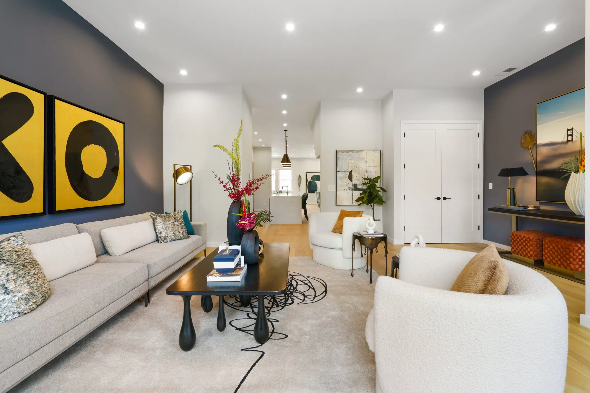 Living room with beige sofa, white armchairs, modern black coffee table, colorful art on navy blue and white walls, decorative vases, and floral arrangements.