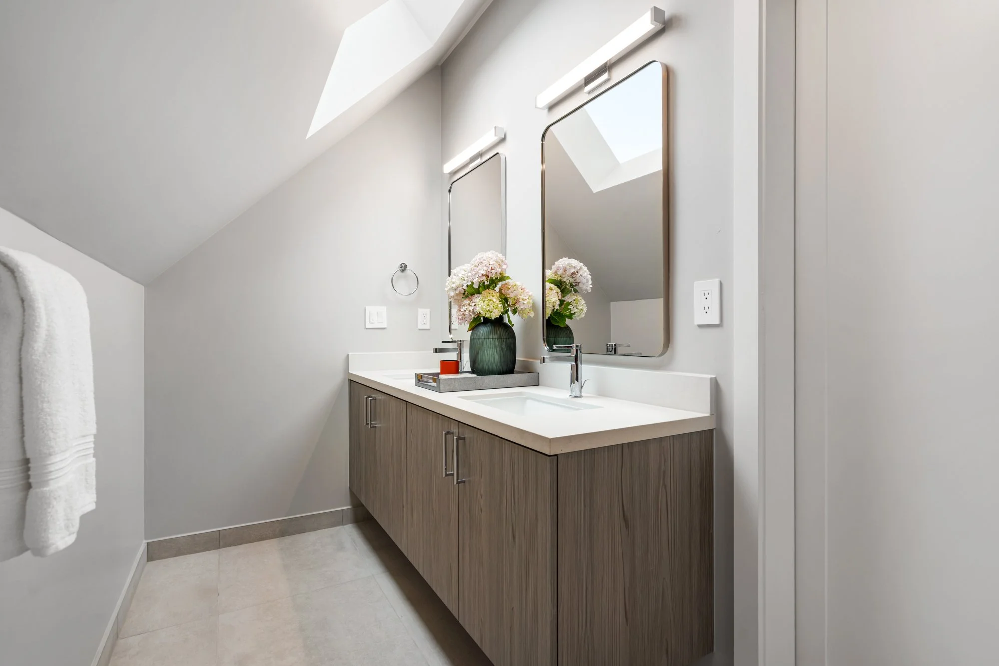 Modern bathroom with light gray walls, a skylight, a wooden vanity with two mirrors, floral arrangement, and minimal decor.
