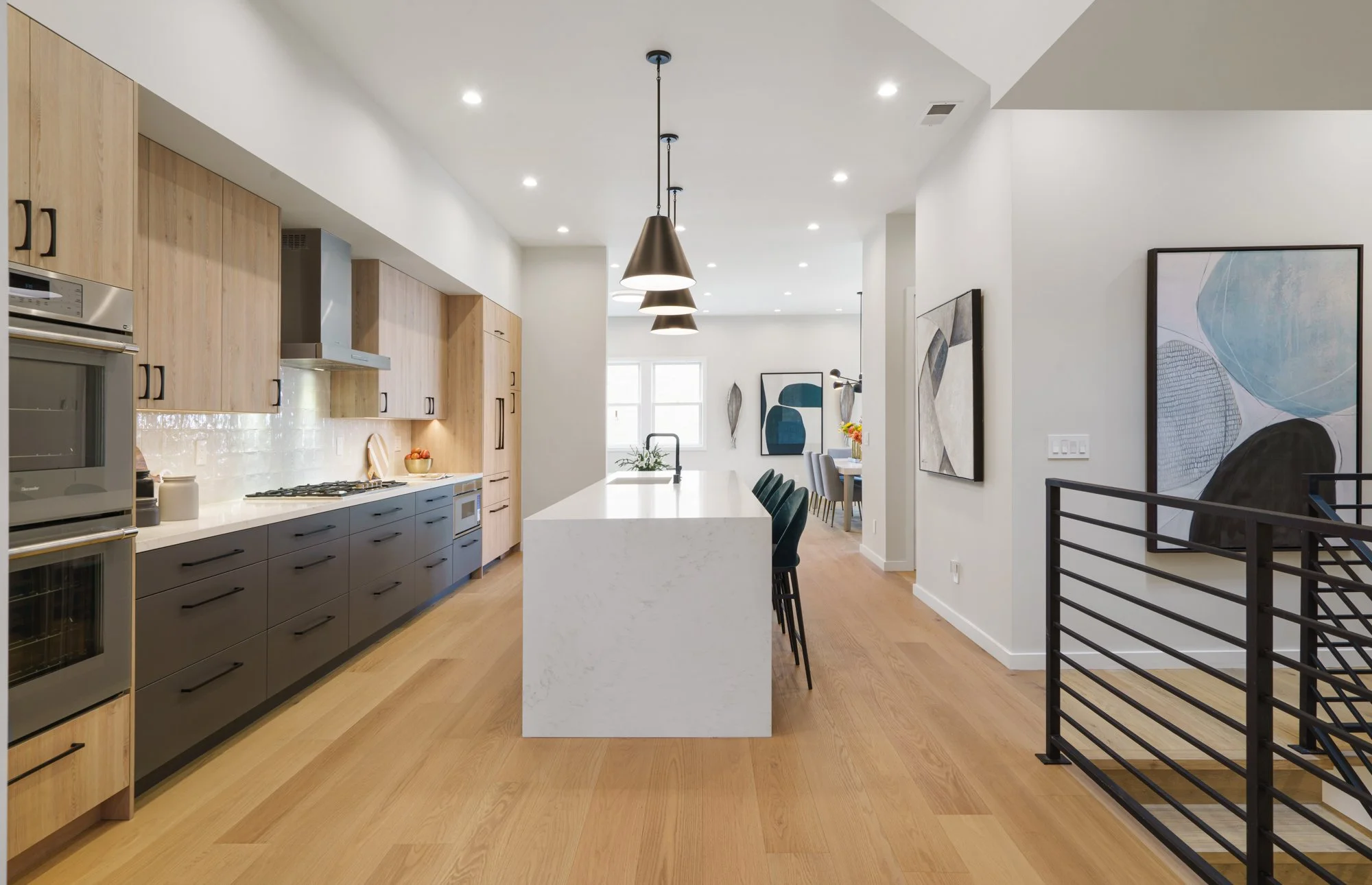 Modern open-concept kitchen with light wood cabinets, black lower cabinets, white island with seating, black pendant lights, and artwork on the walls.