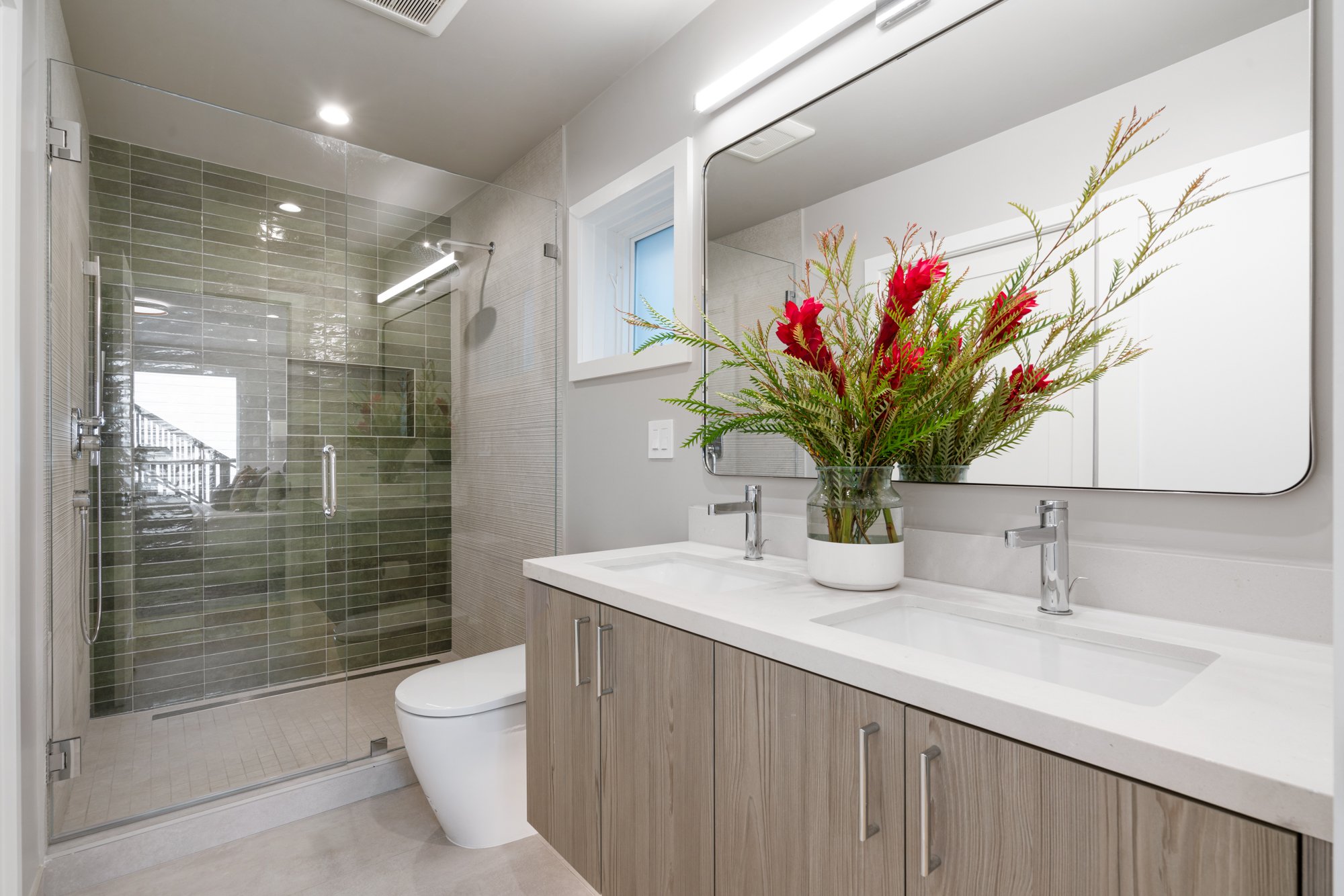 Modern bathroom with dual sinks, a large mirror, a vase with red and green floral arrangement, and a glass-enclosed shower with beige tiles.