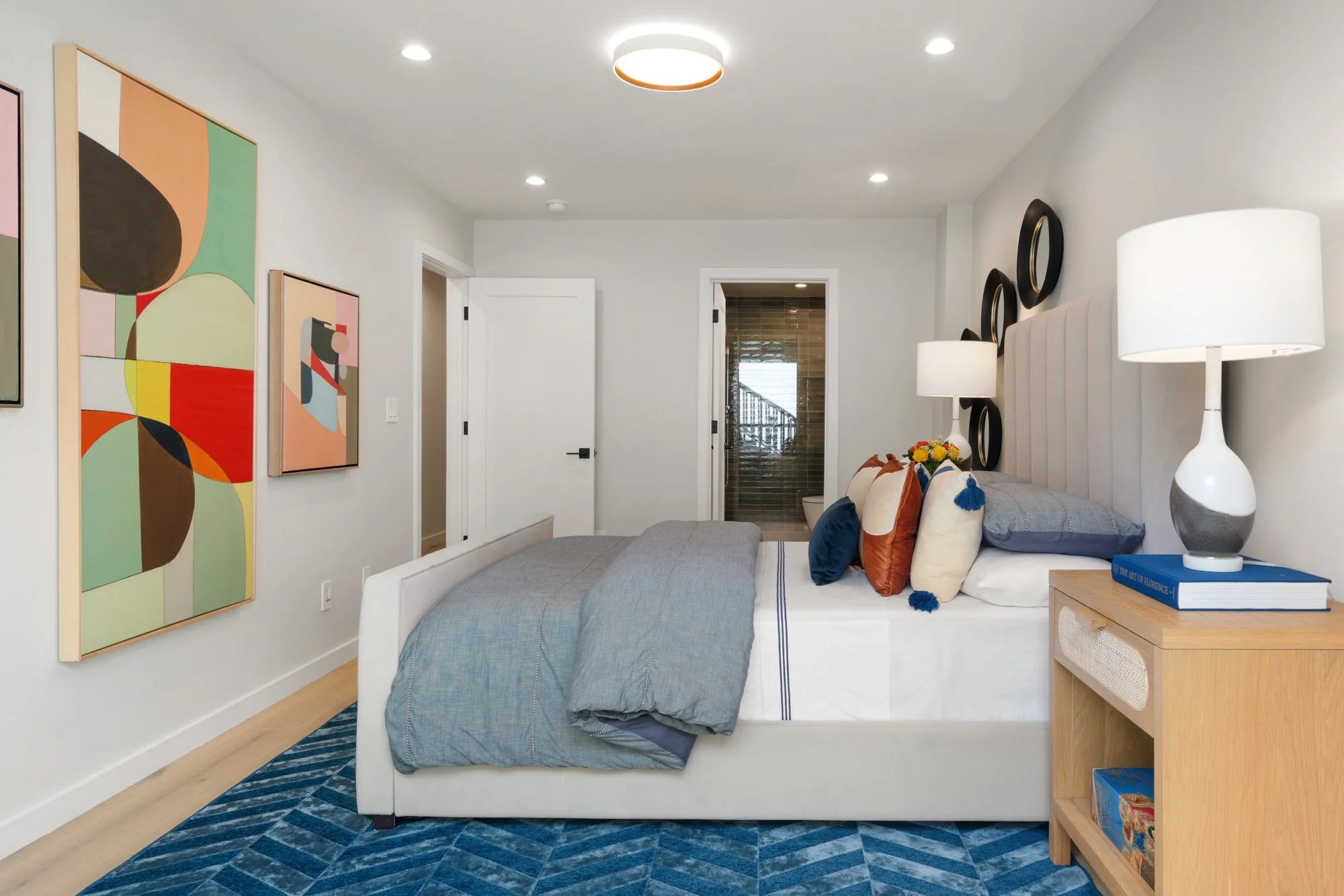 Modern bedroom with white walls, colorful abstract artwork, a white upholstered bed with gray bedding, decorative pillows, a wooden nightstand with a white lamp, and a blue patterned rug.