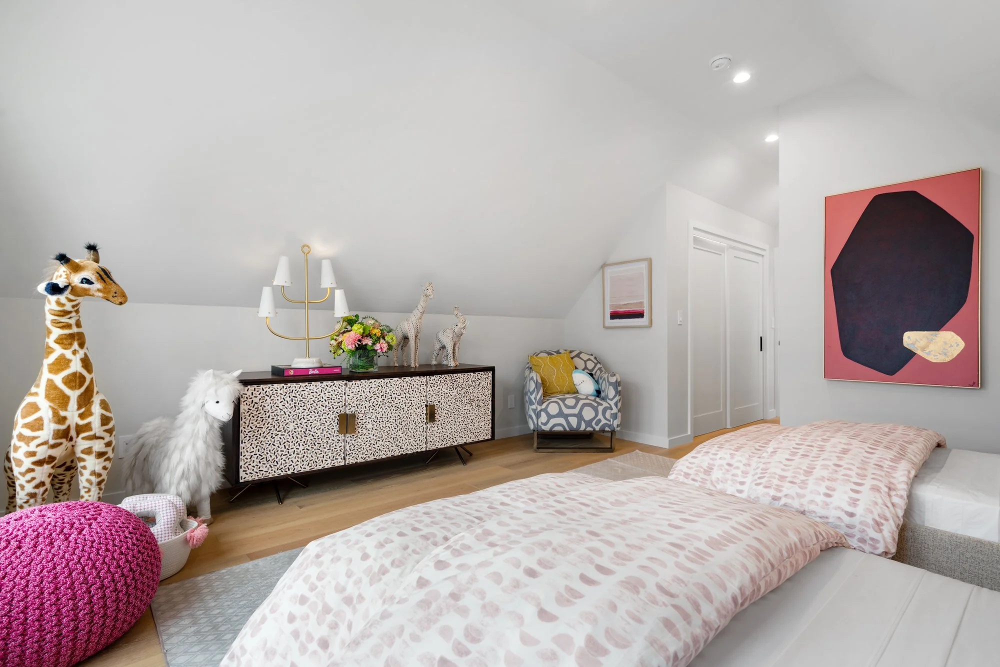 A cozy bedroom with a sloped ceiling, featuring a bed with pink patterned bedding, a white and gray armchair with a yellow pillow, and a modern dresser decorated with books, plants, and giraffe statues. The room includes a large pink and black abstra