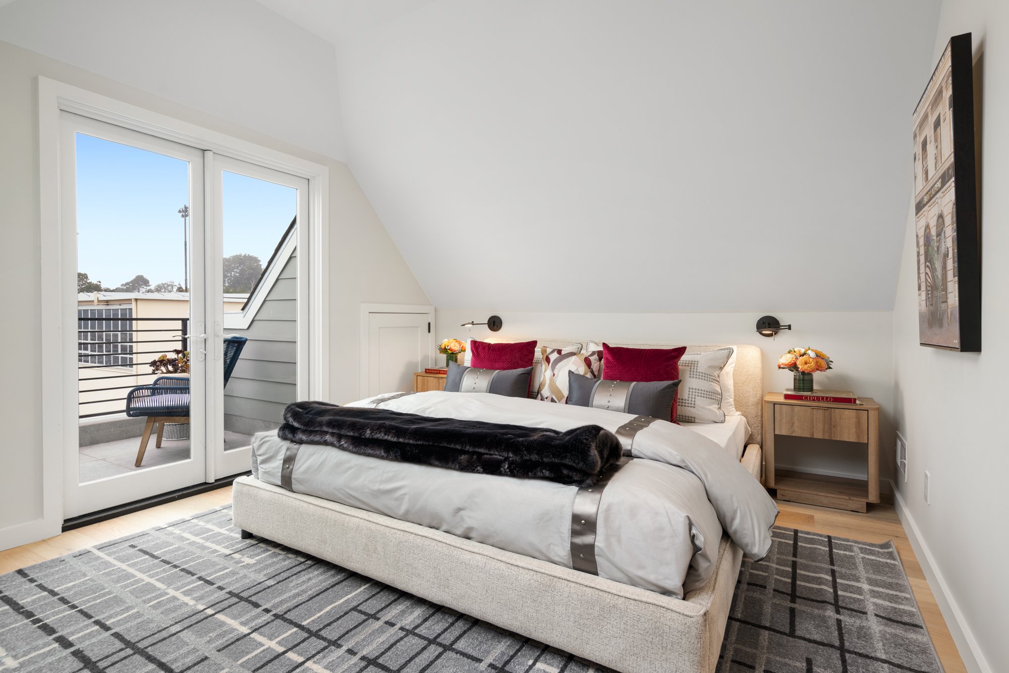 Bedroom with a bed, decorative pillows, bedside tables with flowers, a wall-mounted lamp, a sliding glass door leading to a balcony, a chair outside, and a wall-mounted TV.