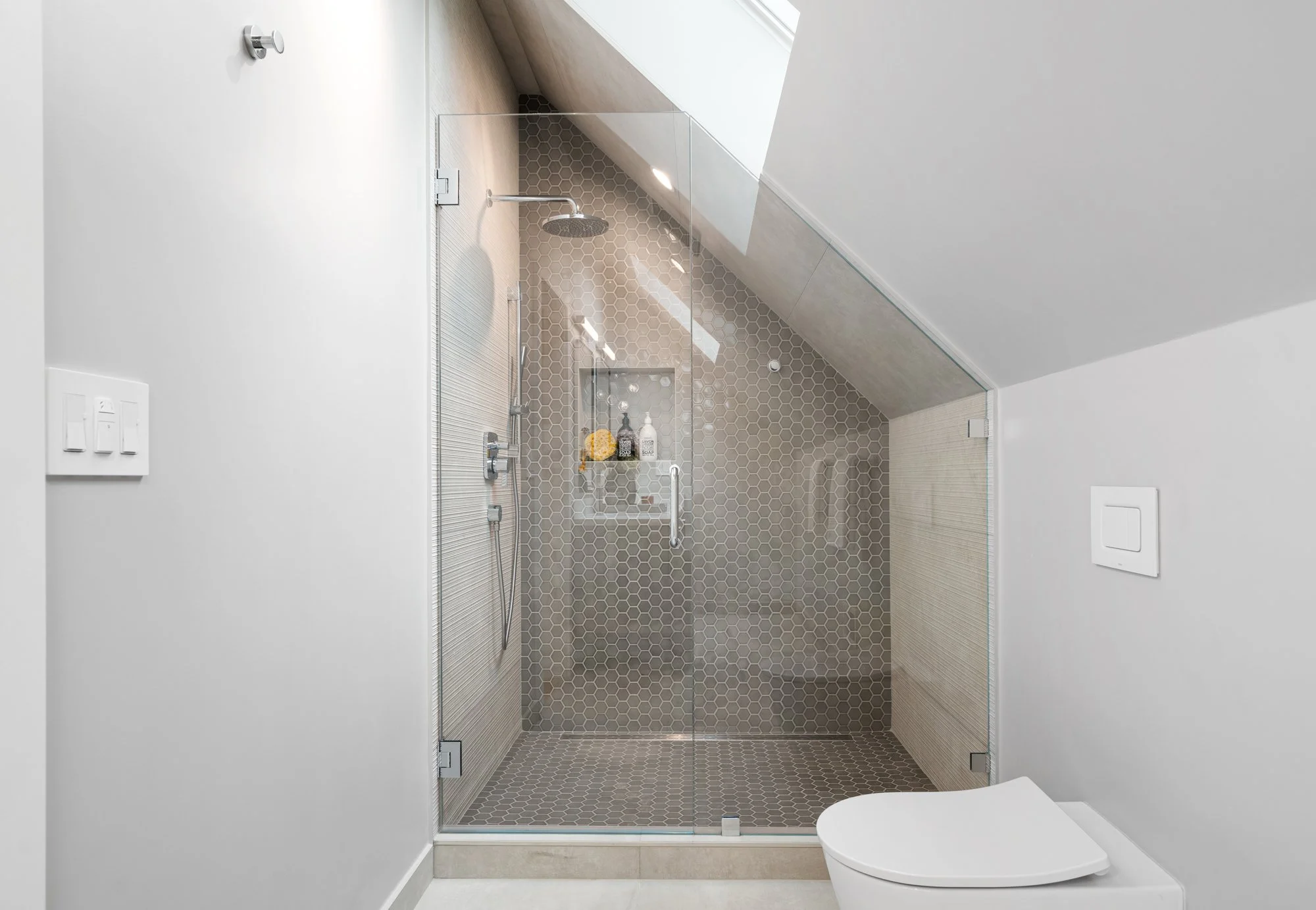Modern bathroom with a glass-enclosed shower area with beige hexagonal tiles on the wall and floor, a skylight, and a white toilet.