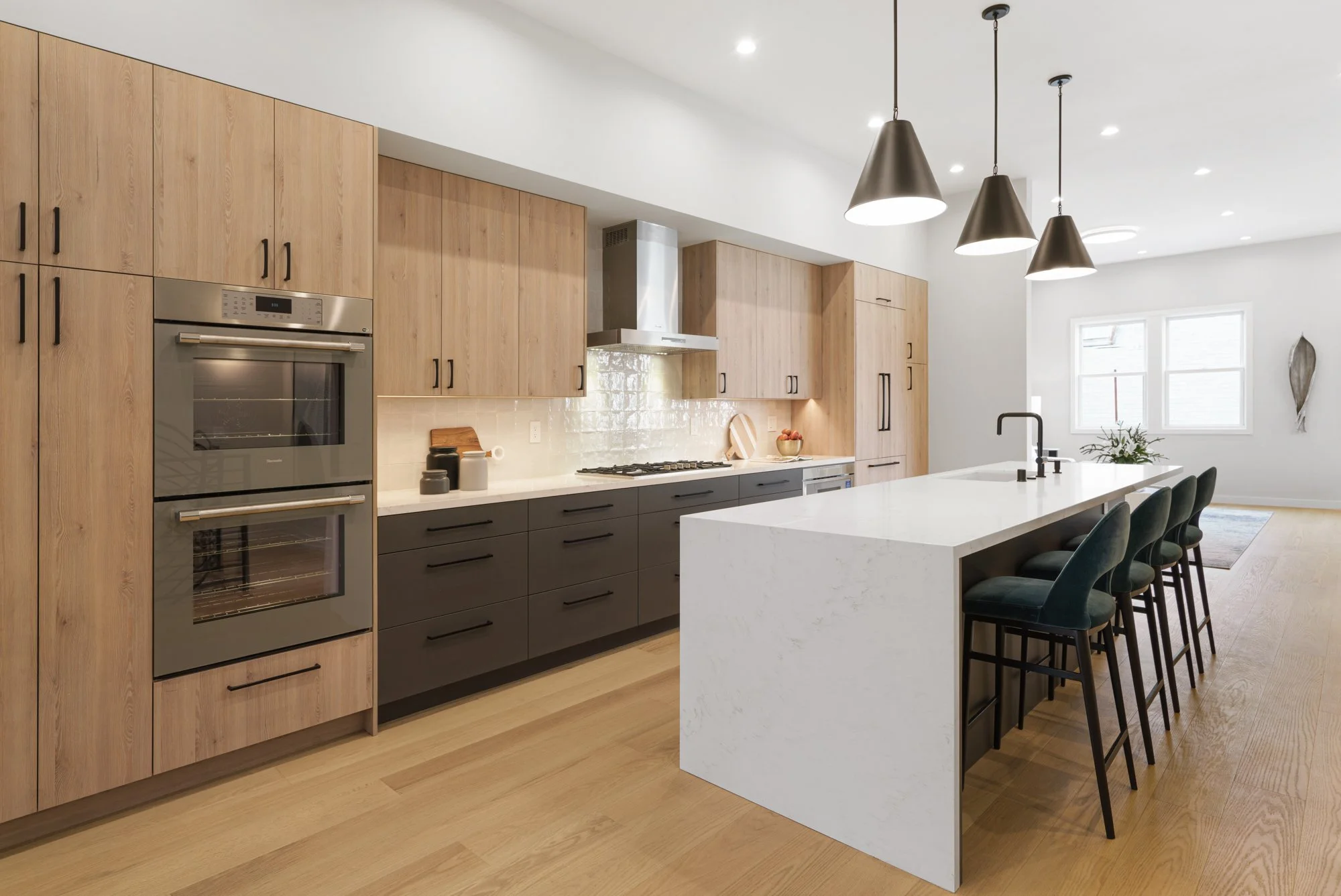 Modern kitchen with wooden cabinets, black lower cabinets, stainless steel oven, white marble island with five black chairs, pendant lights, and natural light from windows.