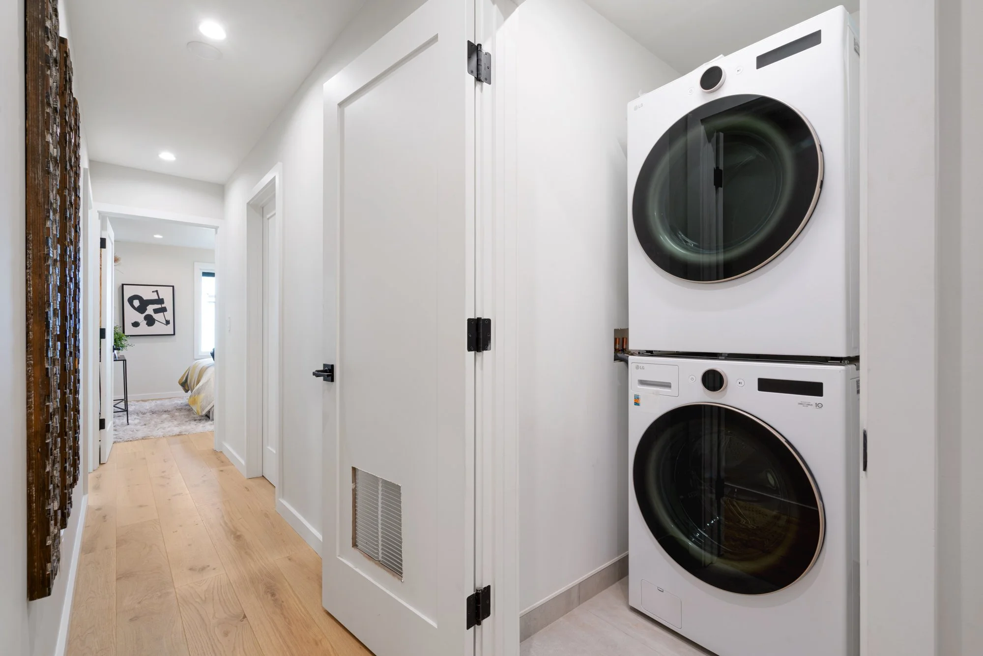 A laundry area with stacked washer and dryer units in a small closet near a hallway leading to a bedroom.