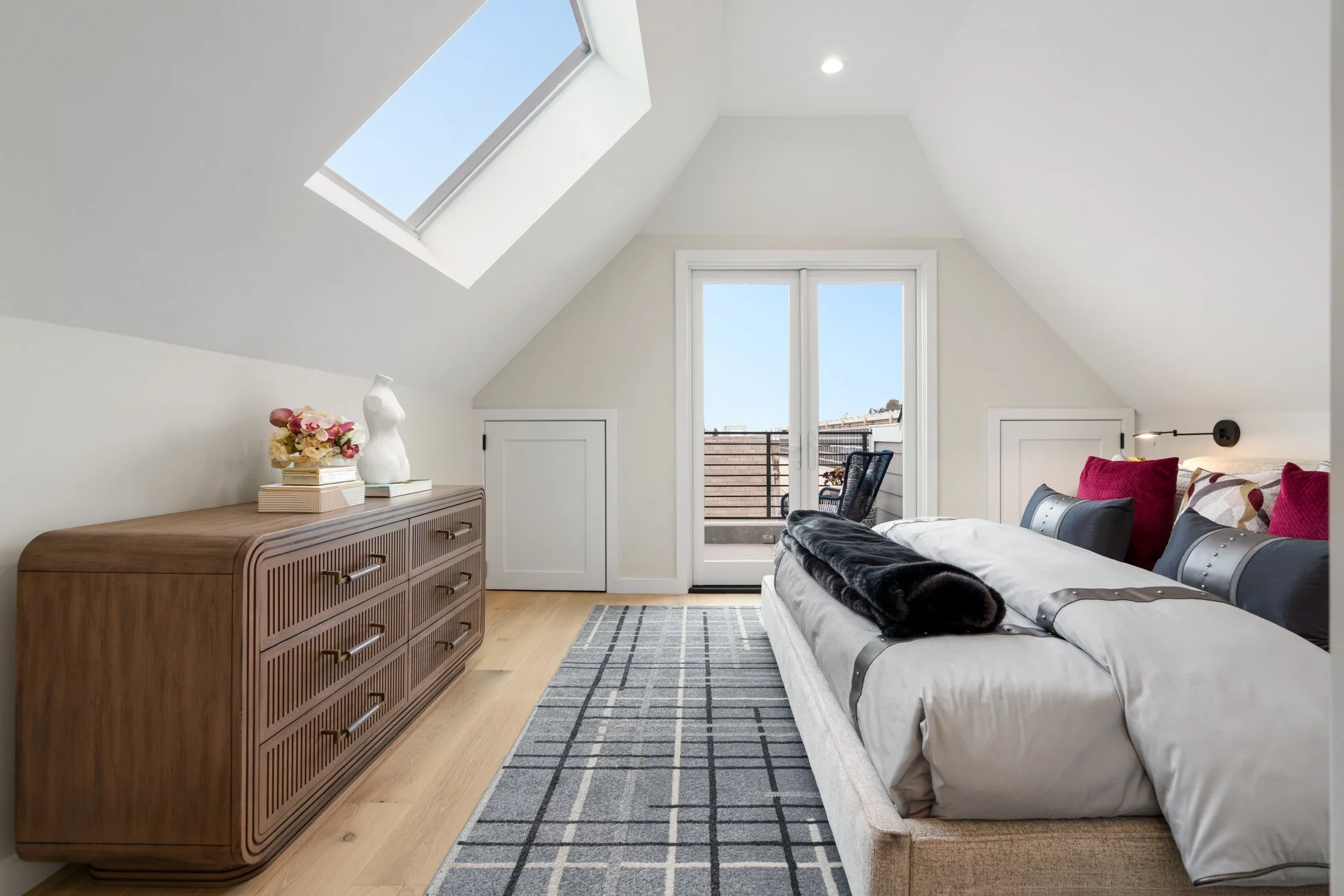 Bright bedroom with sloped ceiling and skylight, featuring a wooden dresser with decorative items, a bed with cushions, and a balcony door letting in natural light.