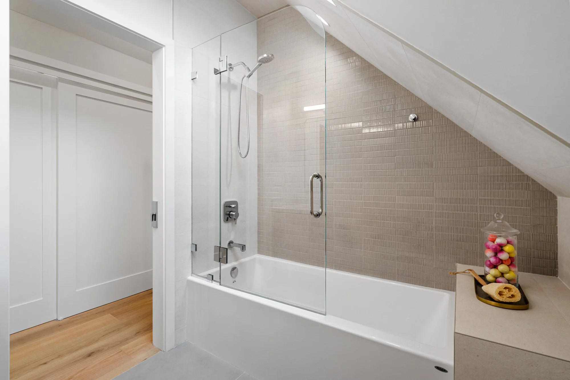 Modern bathroom with white bathtub, clear glass shower door, beige tiled wall, and a jar of colorful decorative eggs on a side surface.