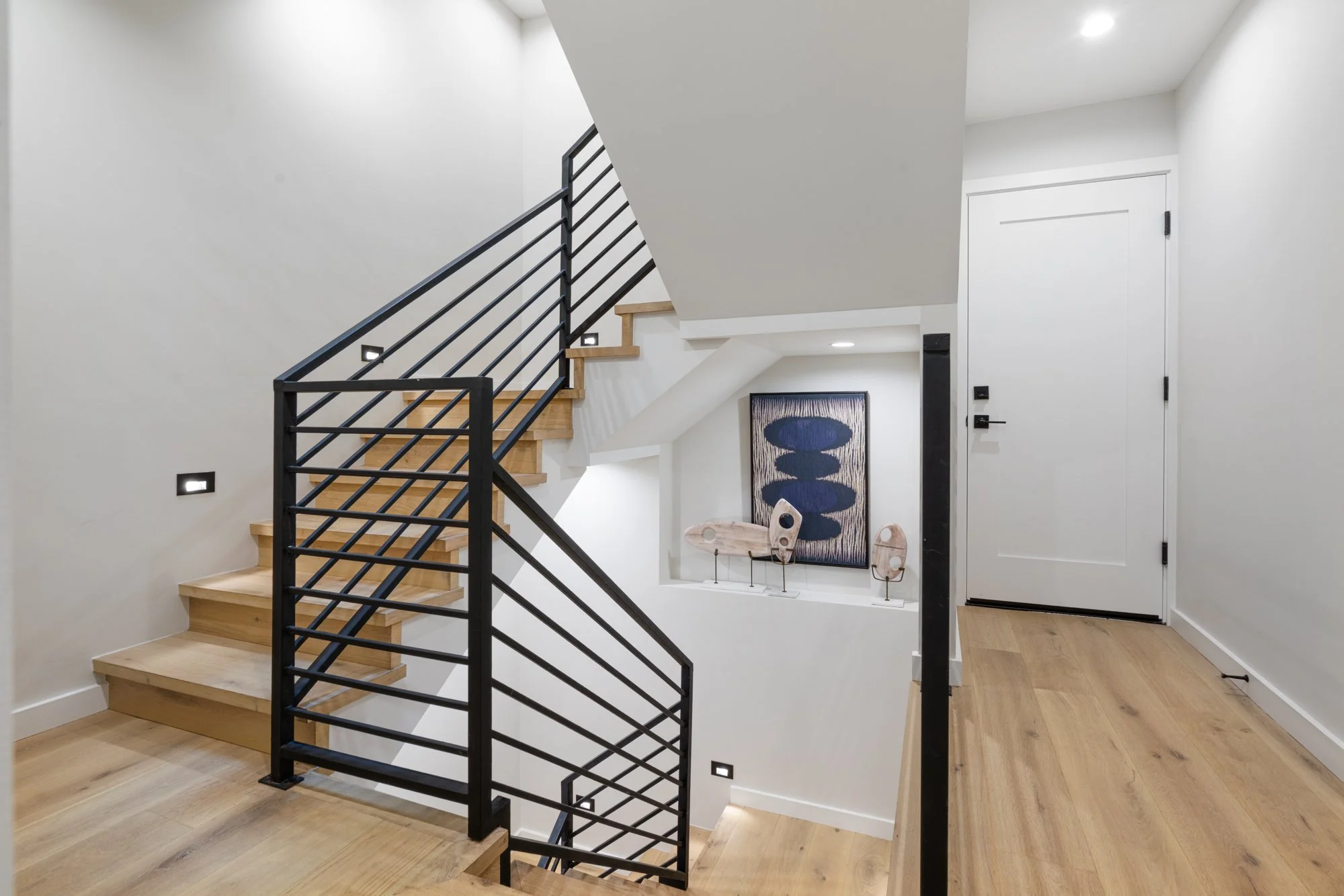 Modern staircase with wooden steps and black metal railing inside a house. White walls, wood flooring, framed artwork, and decorative objects visible in the background.