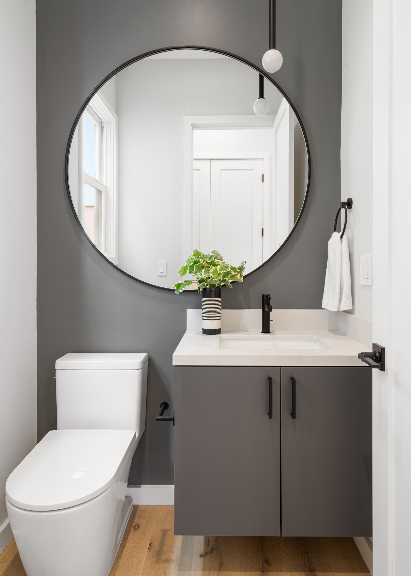 Modern bathroom with a round mirror above a gray vanity, a white toilet, wooden floor, and a black and white striped plant in a black vase.