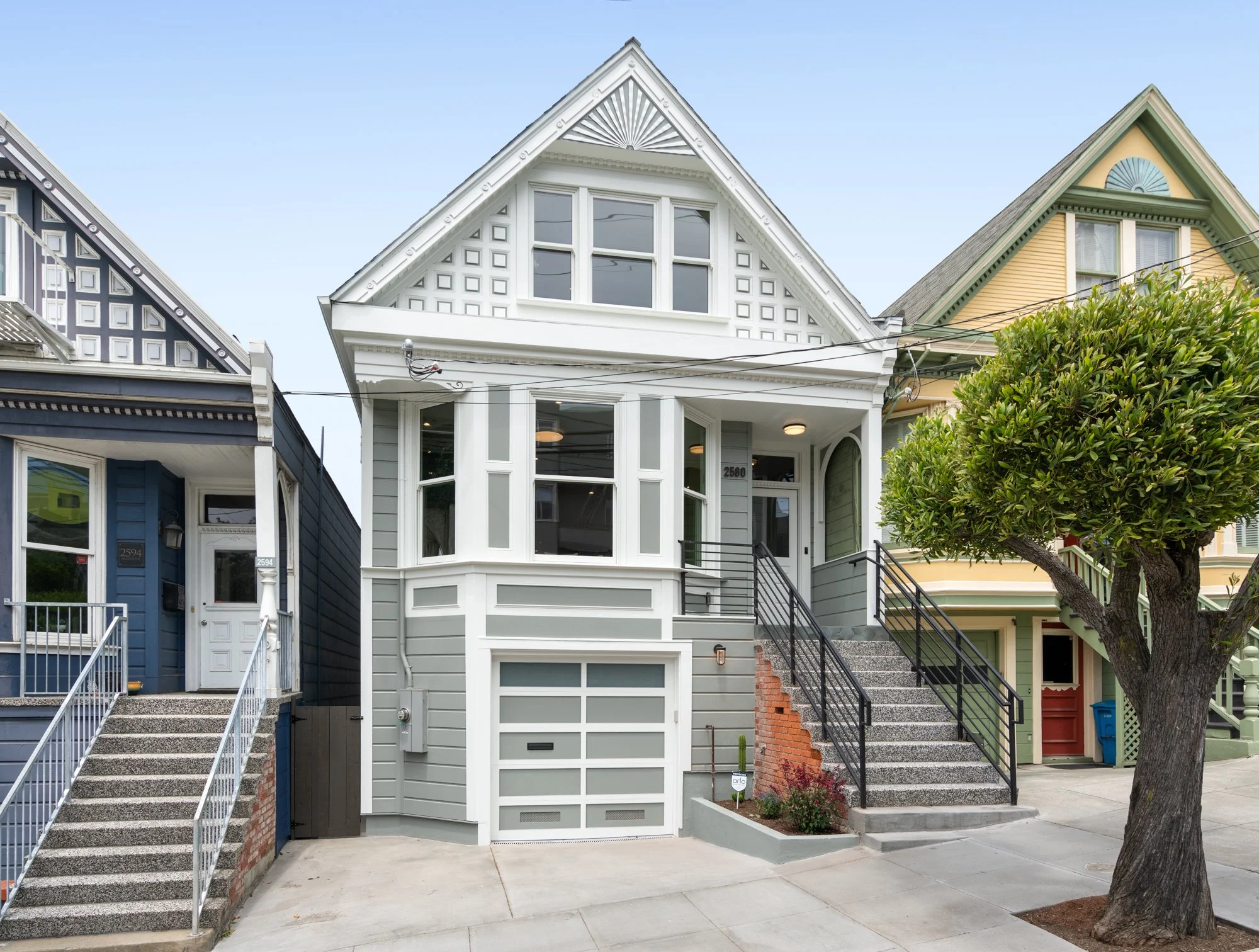Front view of a Victorian style house with a grey exterior, large windows, and front stairs, flanked by neighboring colorful houses and a tree in the sidewalk.