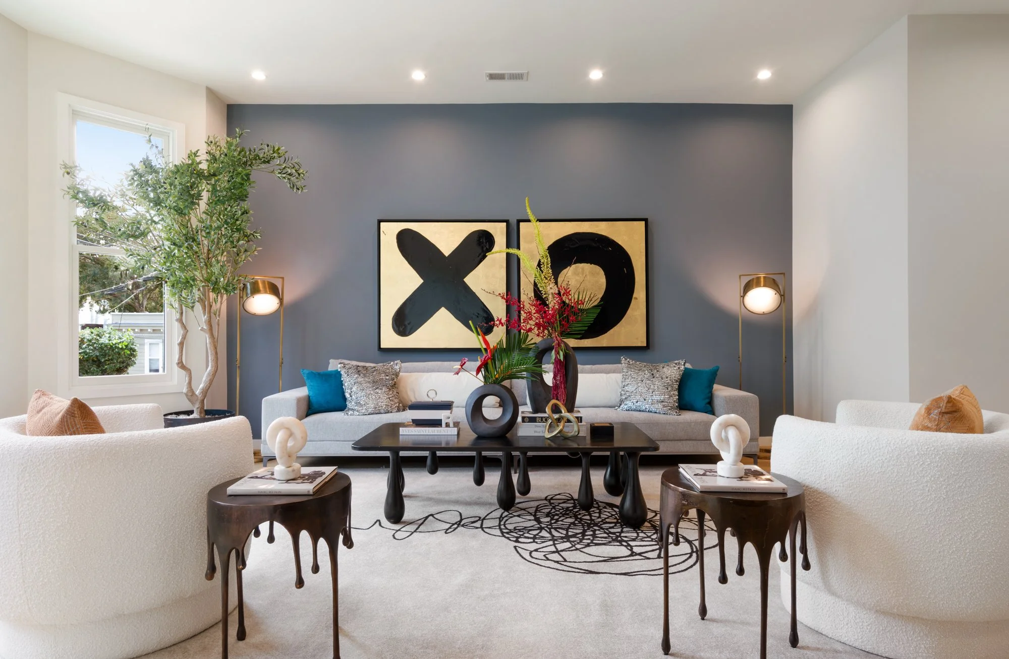 Modern living room with a dark blue accent wall, abstract XO and O artwork, white sofas with colorful pillows, dark wooden side tables with sculptures, a black central table with decorative vases and flowers, large window with greenery outside, moder