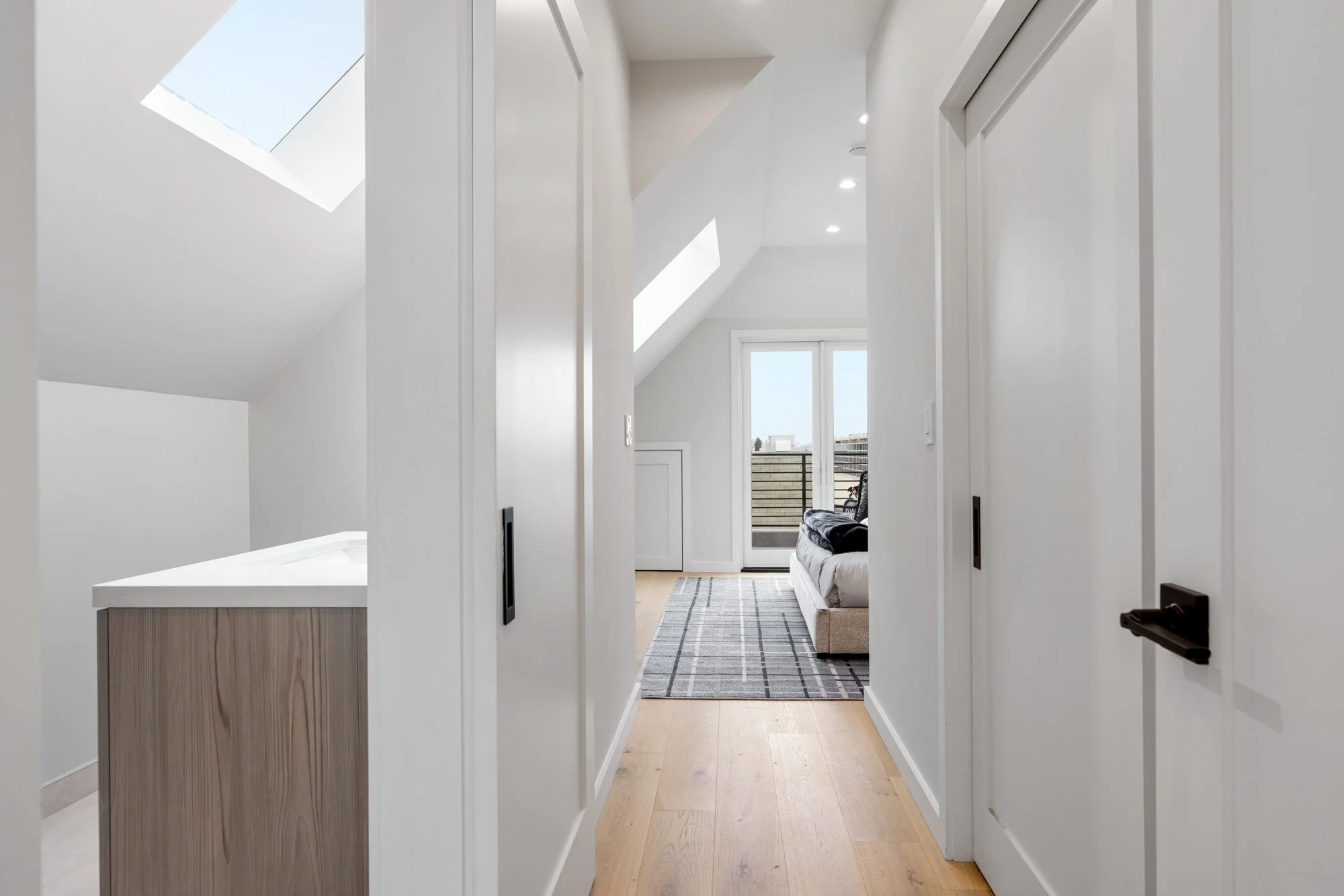 A bright, modern apartment hallway with wooden flooring, white walls, a skylight, and a view of a living room with a glass door leading to a balcony.