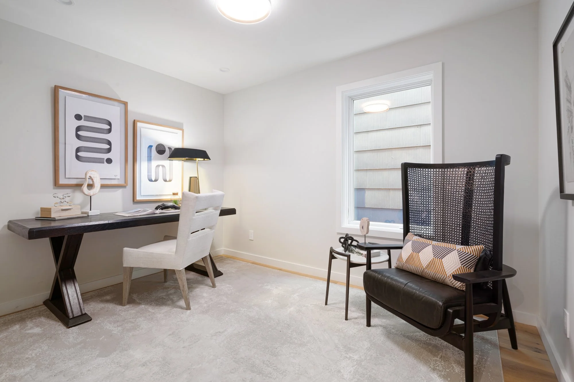 A modern home office with a white desk, an upholstered white chair, black desk lamp, abstract wall art, a black lounge chair with a geometric pillow, and a window with outside siding visible.