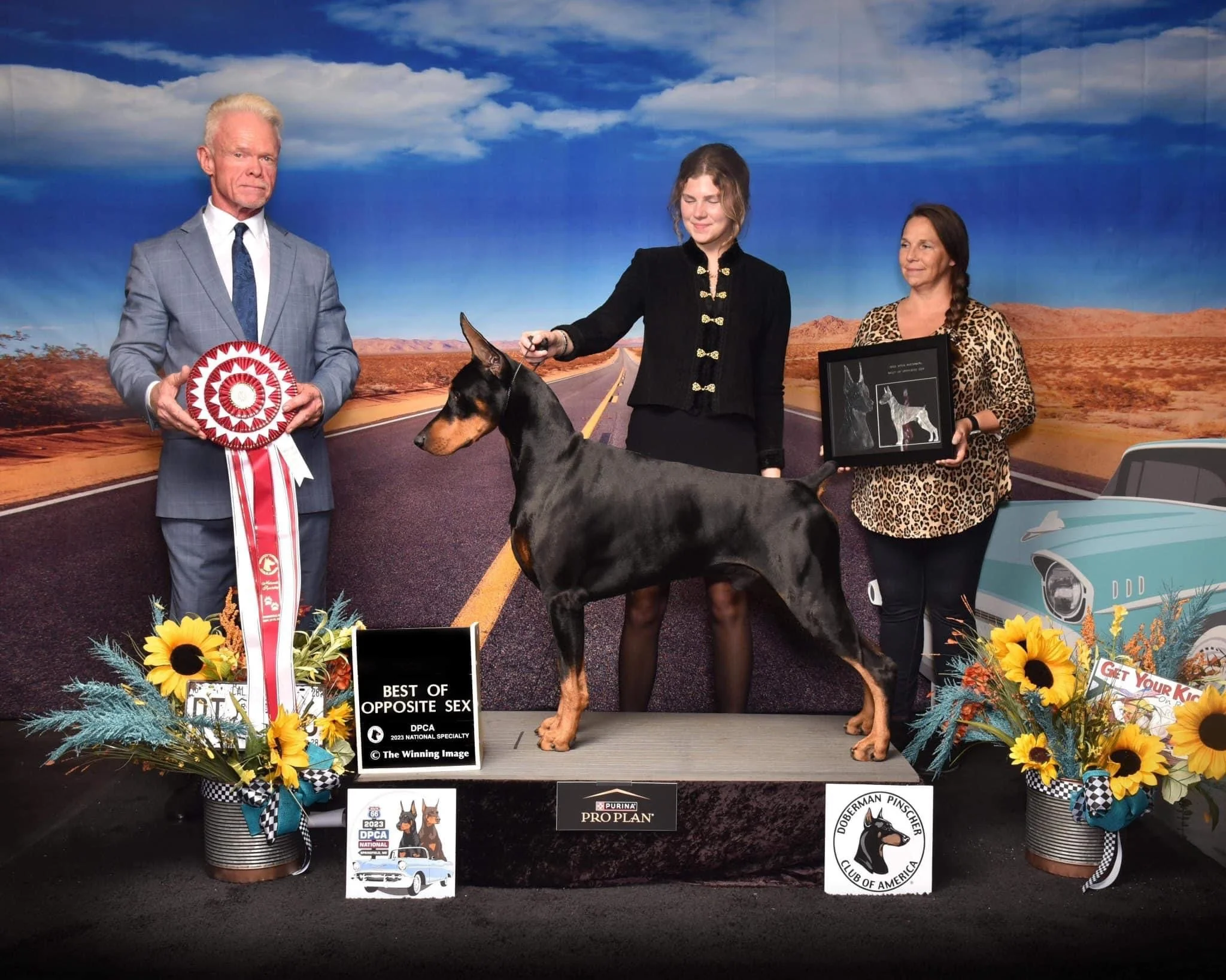 A Doberman Pinscher at a dog show with a handler and judges holding a rosette and plaque, landscape backdrop, floral arrangements, and a sign reading 'Best of Opposite Sex.'