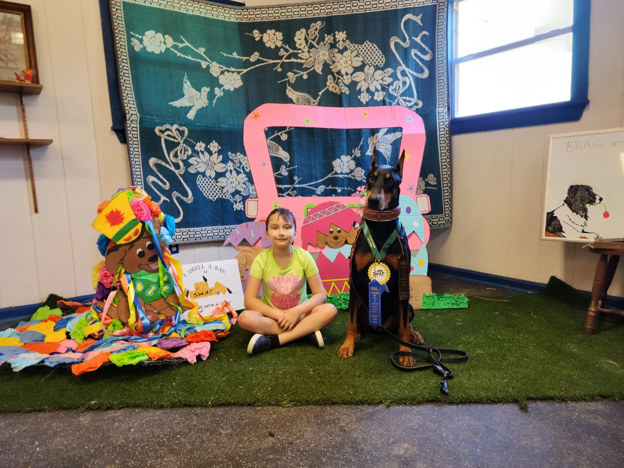 A child sitting on a patch of artificial grass indoors next to a Doberman wearing a blue award ribbon and a medal. The background has colorful decorations including a tapestry with bird and floral designs, paper cutouts, and a cartoon dog sign. A small easel with a dog's illustration is also visible.
