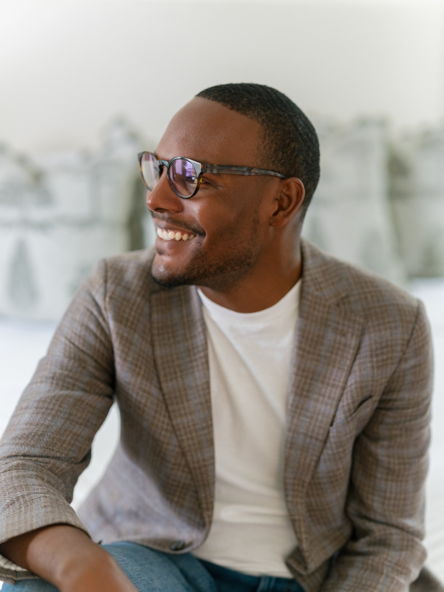 A smiling man wearing glasses, a plaid blazer, a white shirt, and blue jeans, sitting indoors with a blurred background.