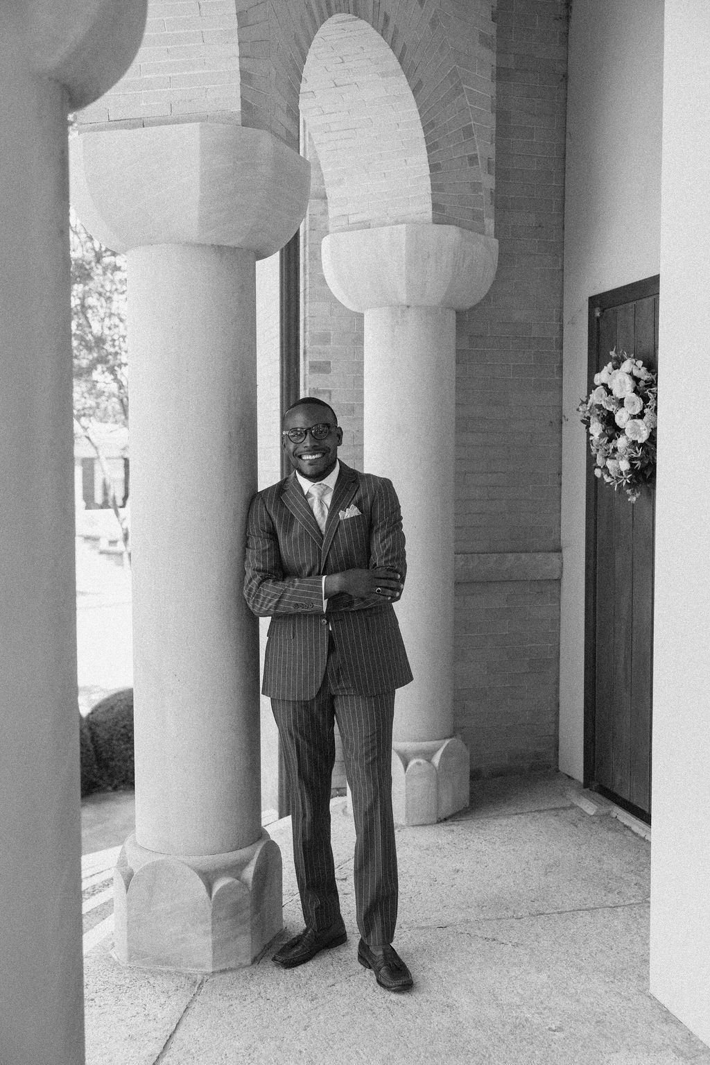 A man in a striped suit with a tie and glasses, smiling and standing with arms crossed, next to a large column, in front of a building with a decorated door and floral wreath.
