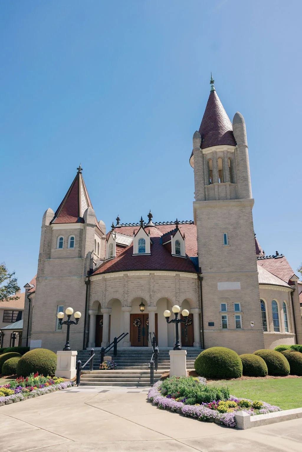 A large castle-like building with turrets, arched windows, and a red-tiled roof, surrounded by a landscaped garden with bushes, flowers, and lamp posts under a clear blue sky.