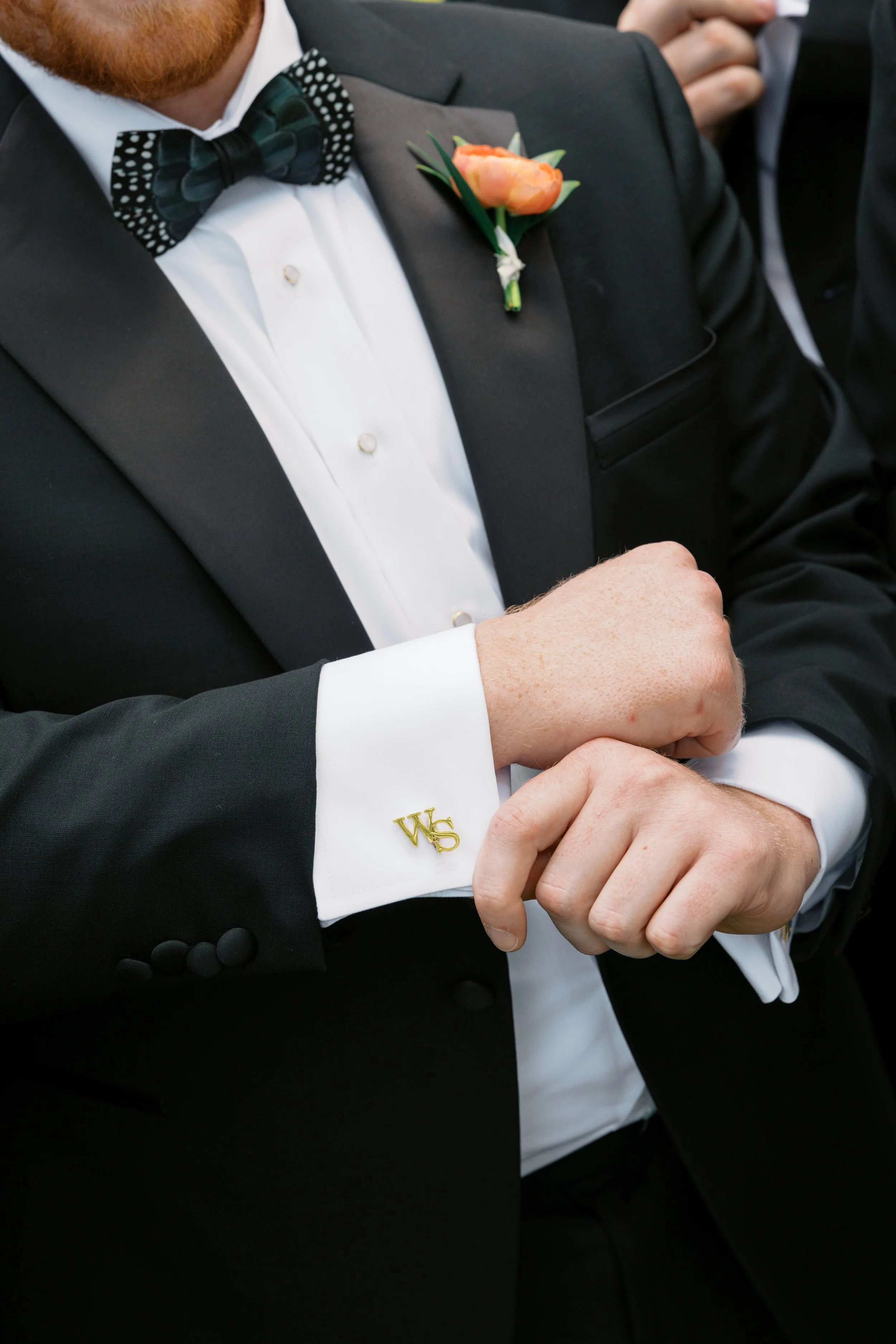Close-up of a man dressed in a black tuxedo with a white shirt, black patterned bow tie, an orange boutonniere on his lapel, and gold cufflinks with initials, at a formal event.