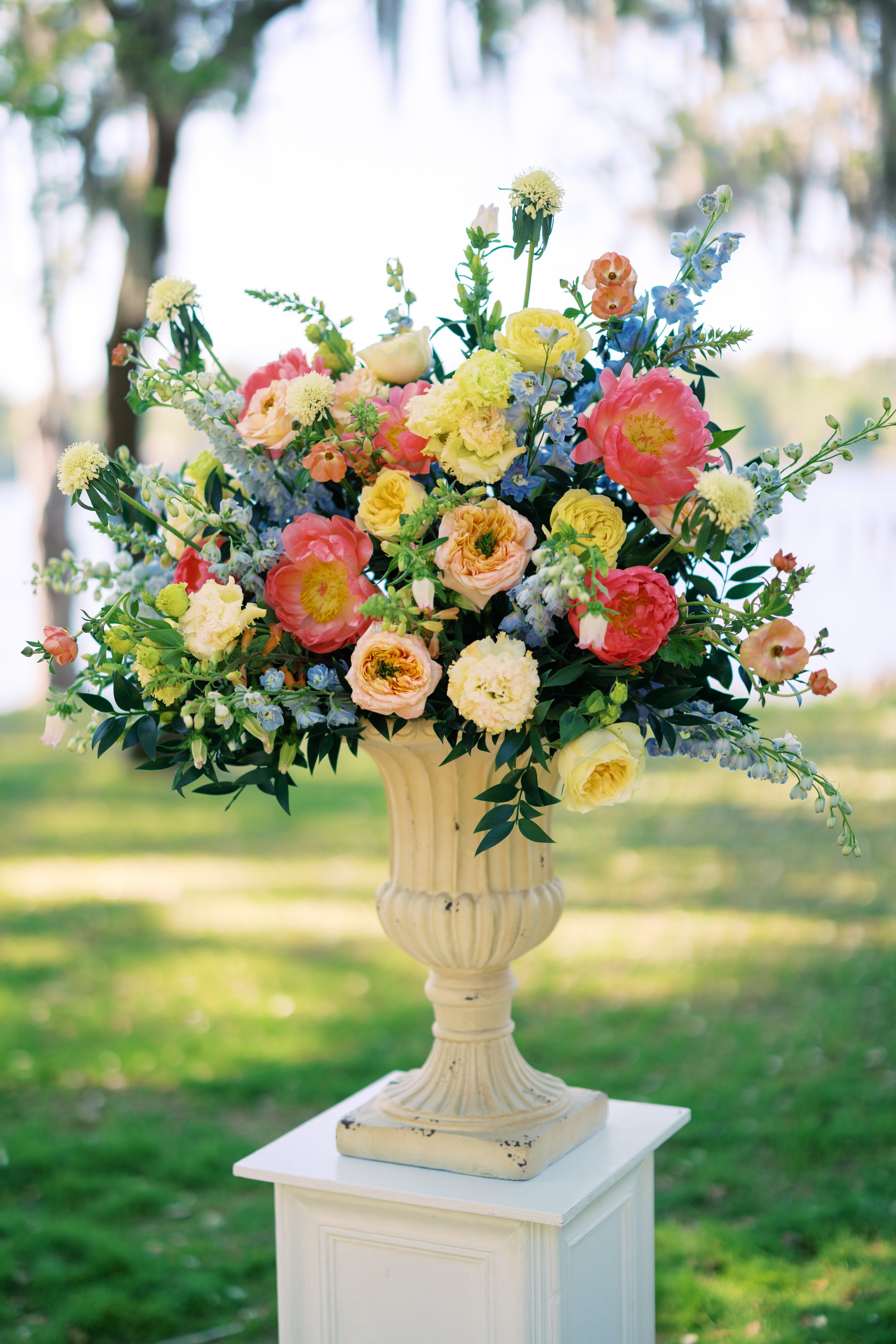 A large bouquet of colorful flowers in a vintage cream-colored pedestal vase on a white table outdoors with trees and grass in the background.