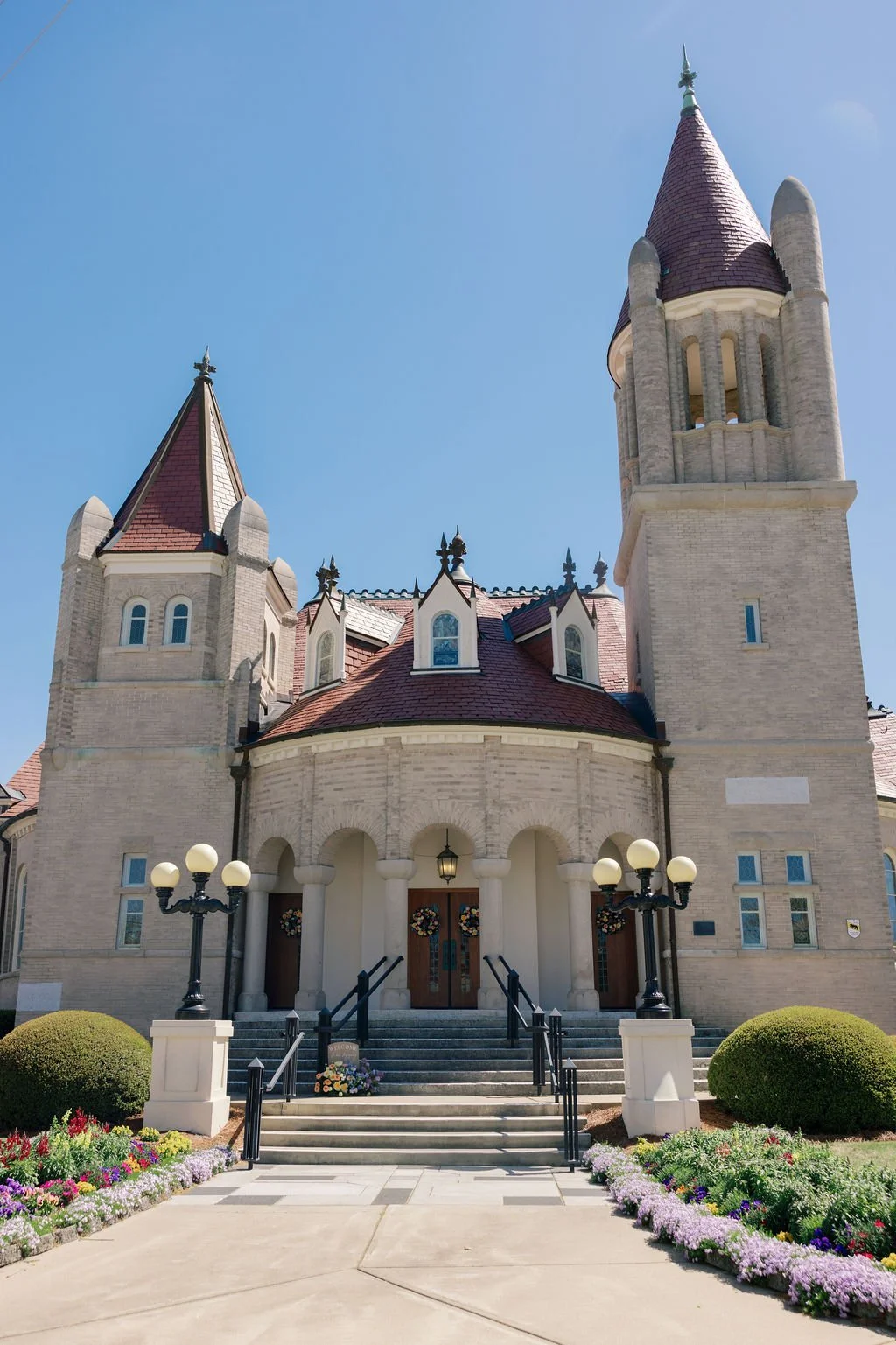 A castle-like building with two tall towers, arched entry with stairs, surrounded by flowering plants and well-manicured bushes, under a clear blue sky.