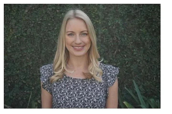 A young woman with long blonde hair smiling, standing outdoors in front of green foliage.