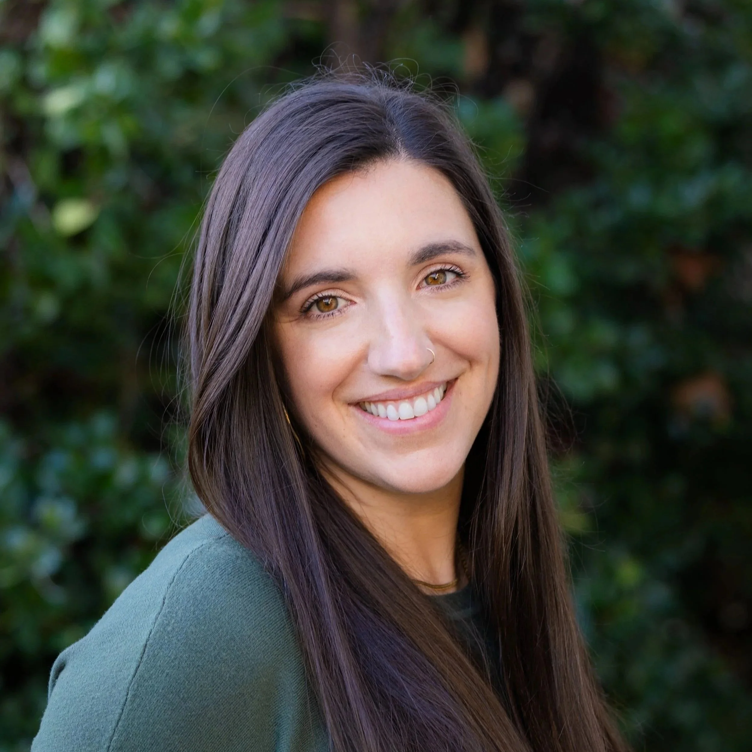 A smiling woman with long brown hair and light brown eyes, wearing a green top, standing outdoors with a blurred green foliage background.