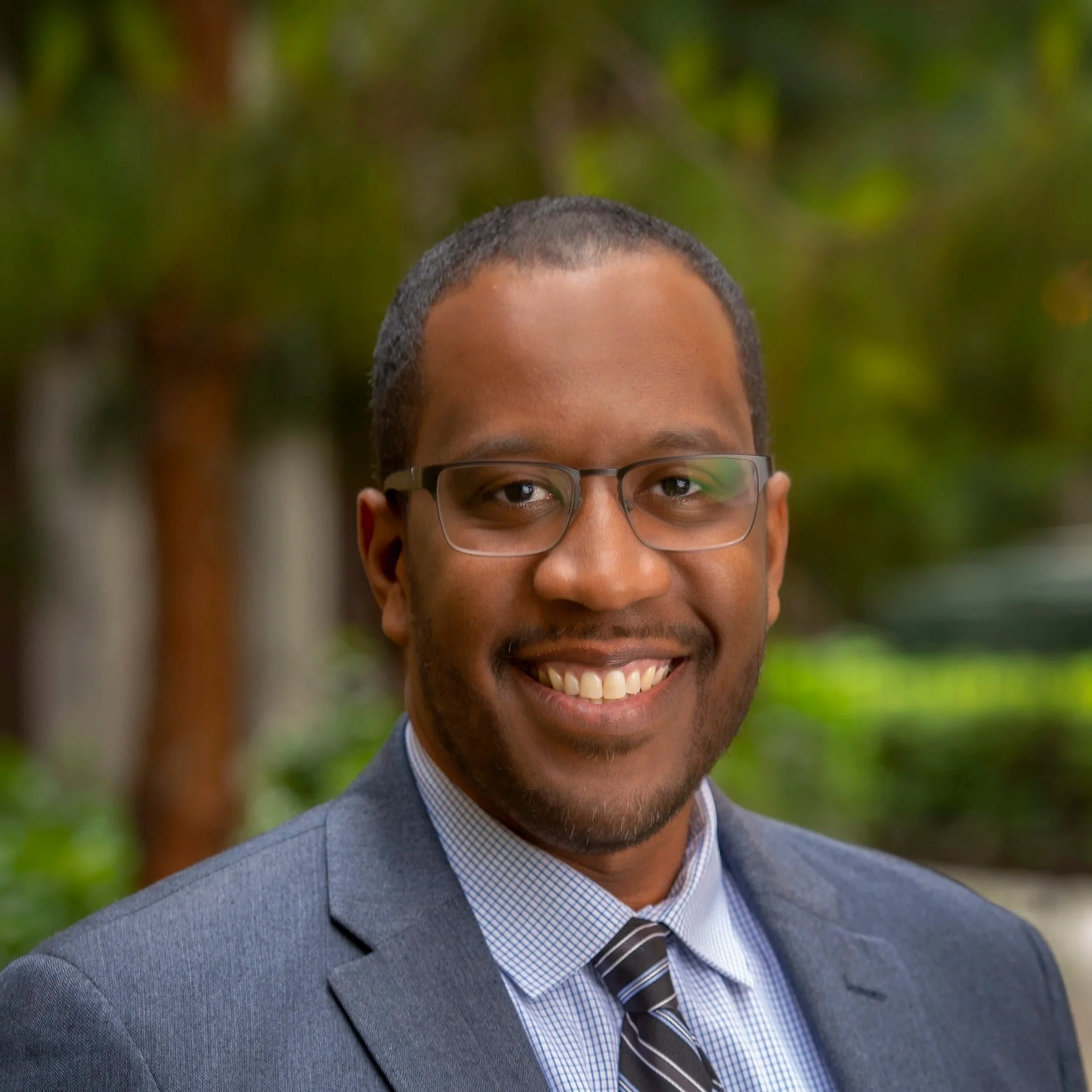 A smiling man wearing glasses, a gray suit, a light blue shirt, and a striped tie outdoors with blurred greenery in the background.