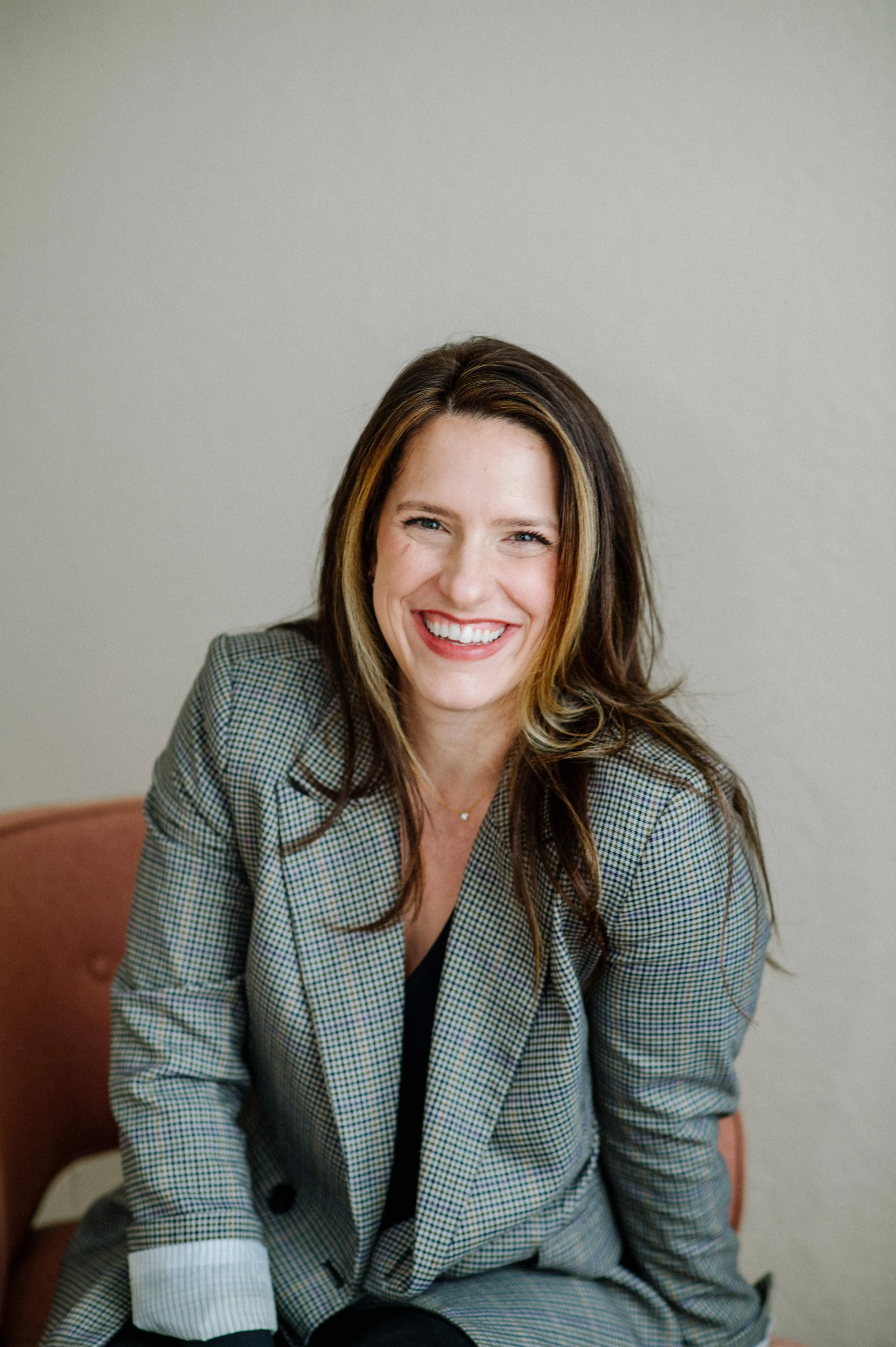A woman with long brown hair and a big smile, wearing a checked blazer, sitting on a pink chair against a plain wall.