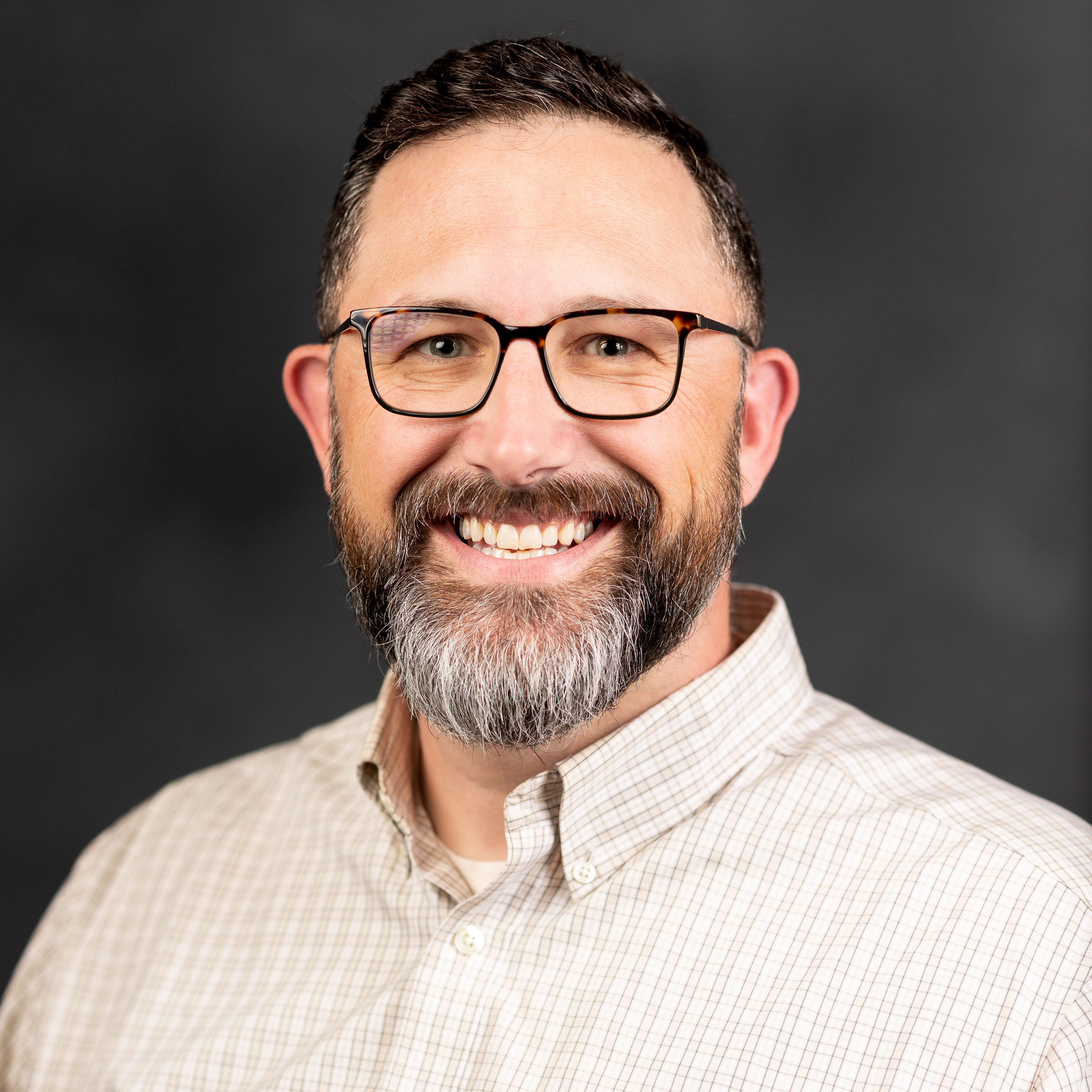 Portrait of a smiling man with glasses, beard, and mustache wearing a beige checkered shirt against a dark background.