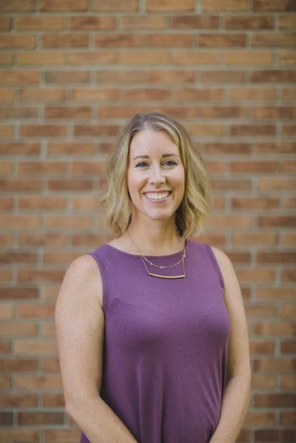 A woman with shoulder-length blonde hair wearing a purple sleeveless top and a simple necklace, smiling in front of a brick wall.