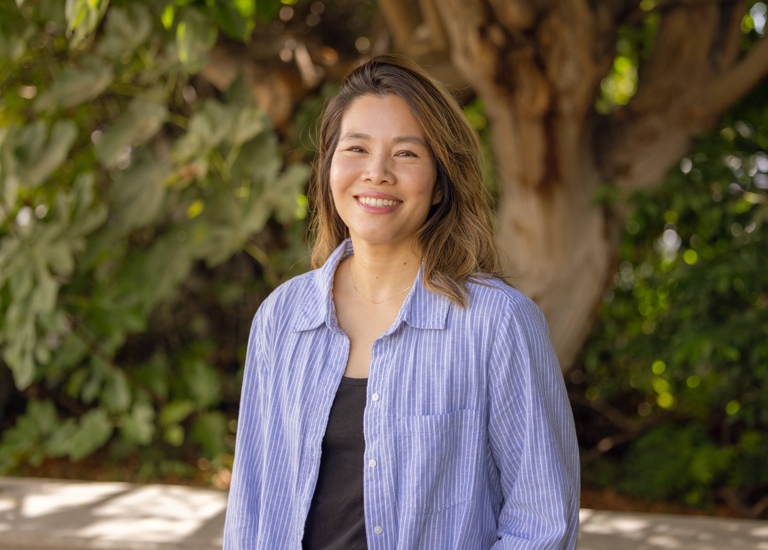 A woman smiling outdoors in front of a tree and green foliage.