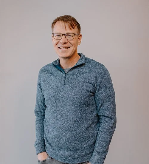 Person wearing glasses and a blue sweater standing against a plain background.