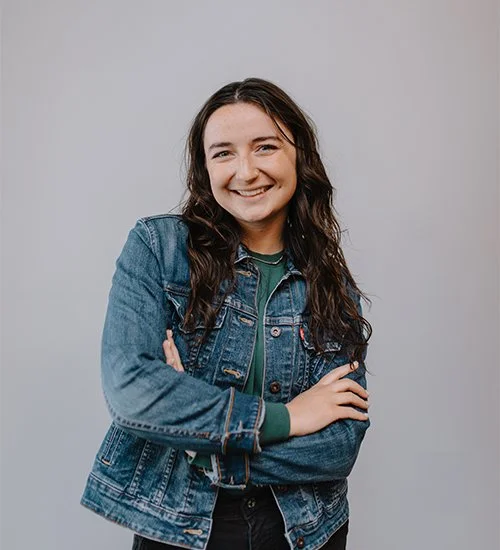 A person with long dark hair smiling and wearing a denim jacket over a green top with arms crossed.